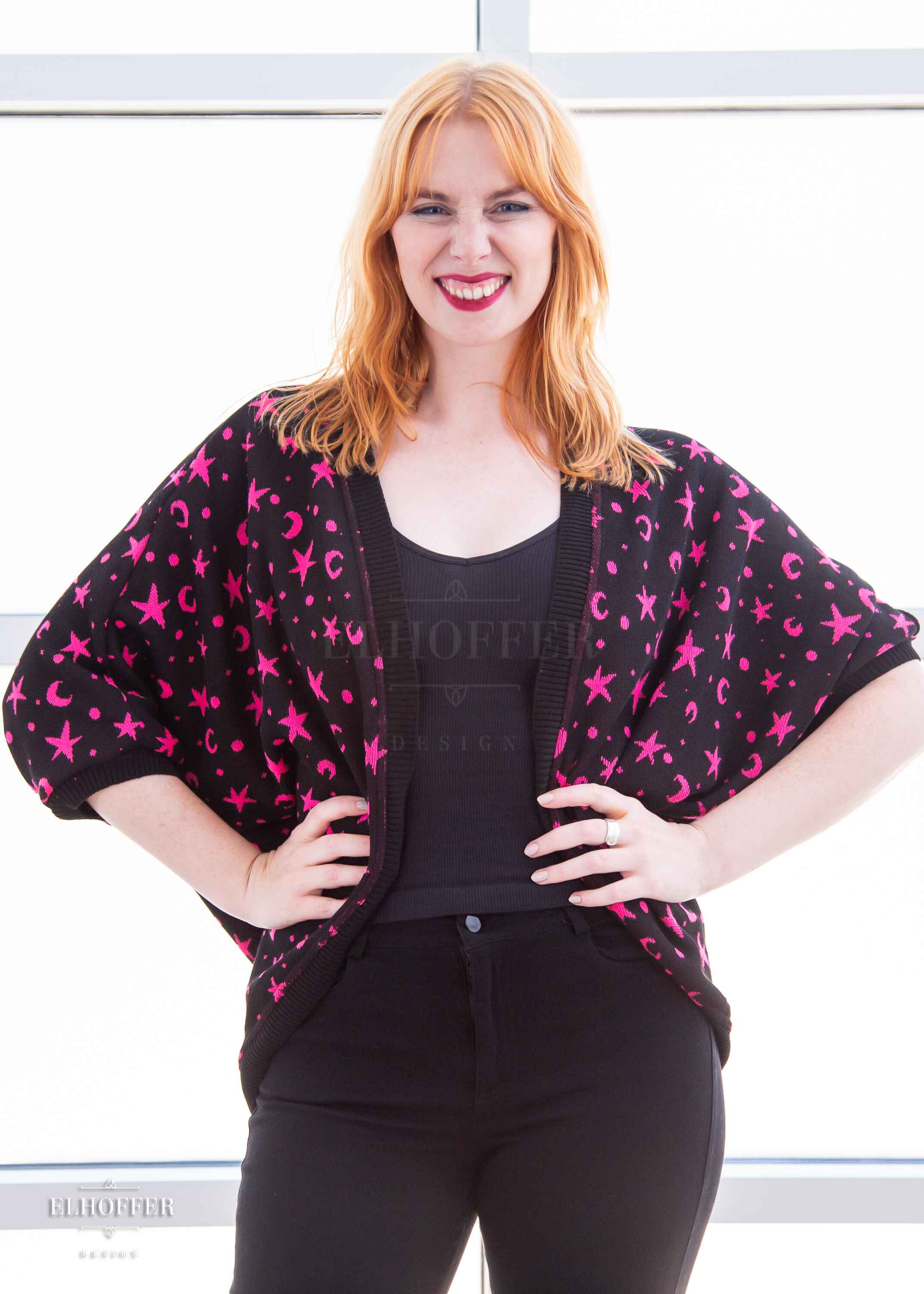 Harley, a fair skinned S model with shoulder length strawberry blonde hair, is smiling while wearing an open front dolman with 3/4 sleeves. The main body of the dolman is black with a bright pink star and moon pattern knit throughout. The edges and cuffs are ribbed in black.