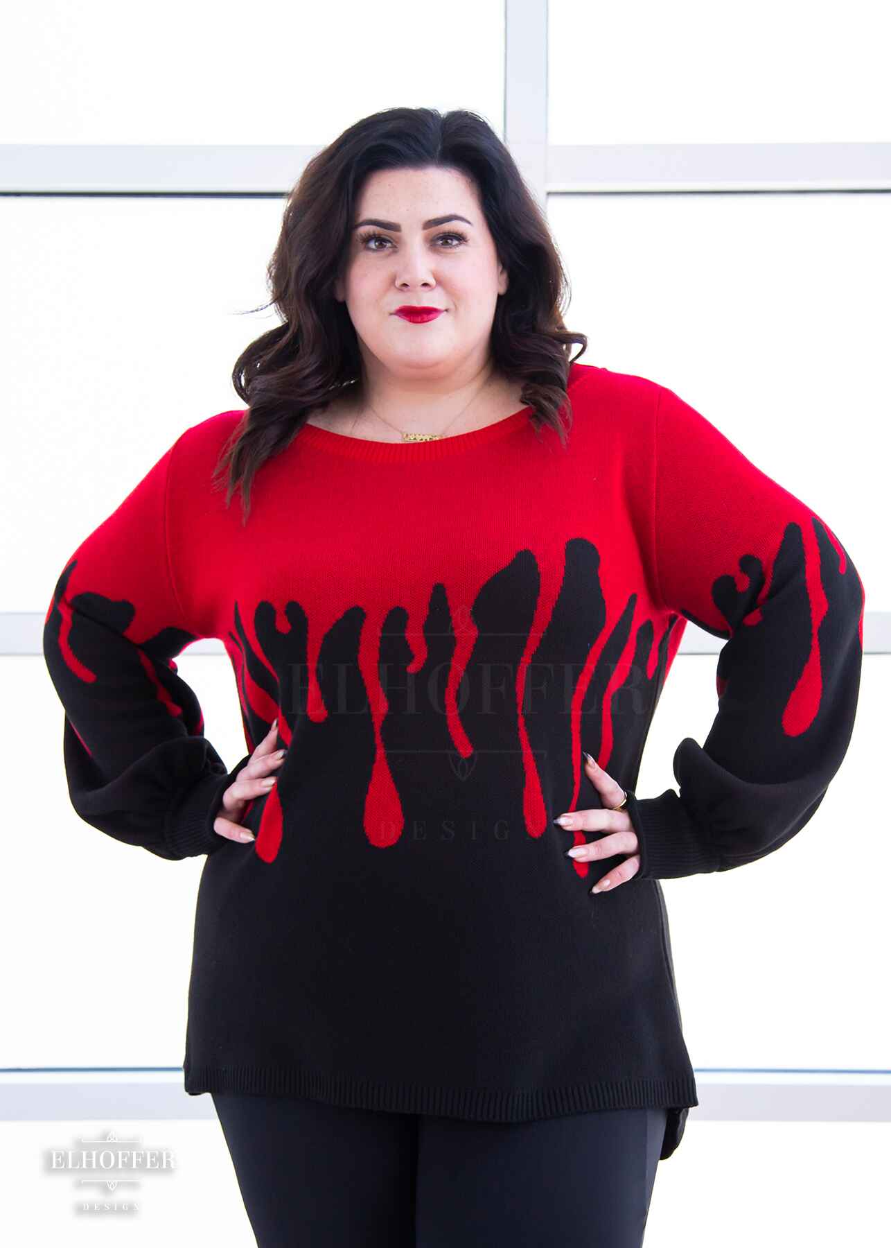 Stacy, a fair skinned XL model with shoulder length wavy dark brown hair, is wearing an oversize sweater with a bright red blood drip design that looks like it's oozing down onto a black sweater that has long billowing sleeves with thumbholes.