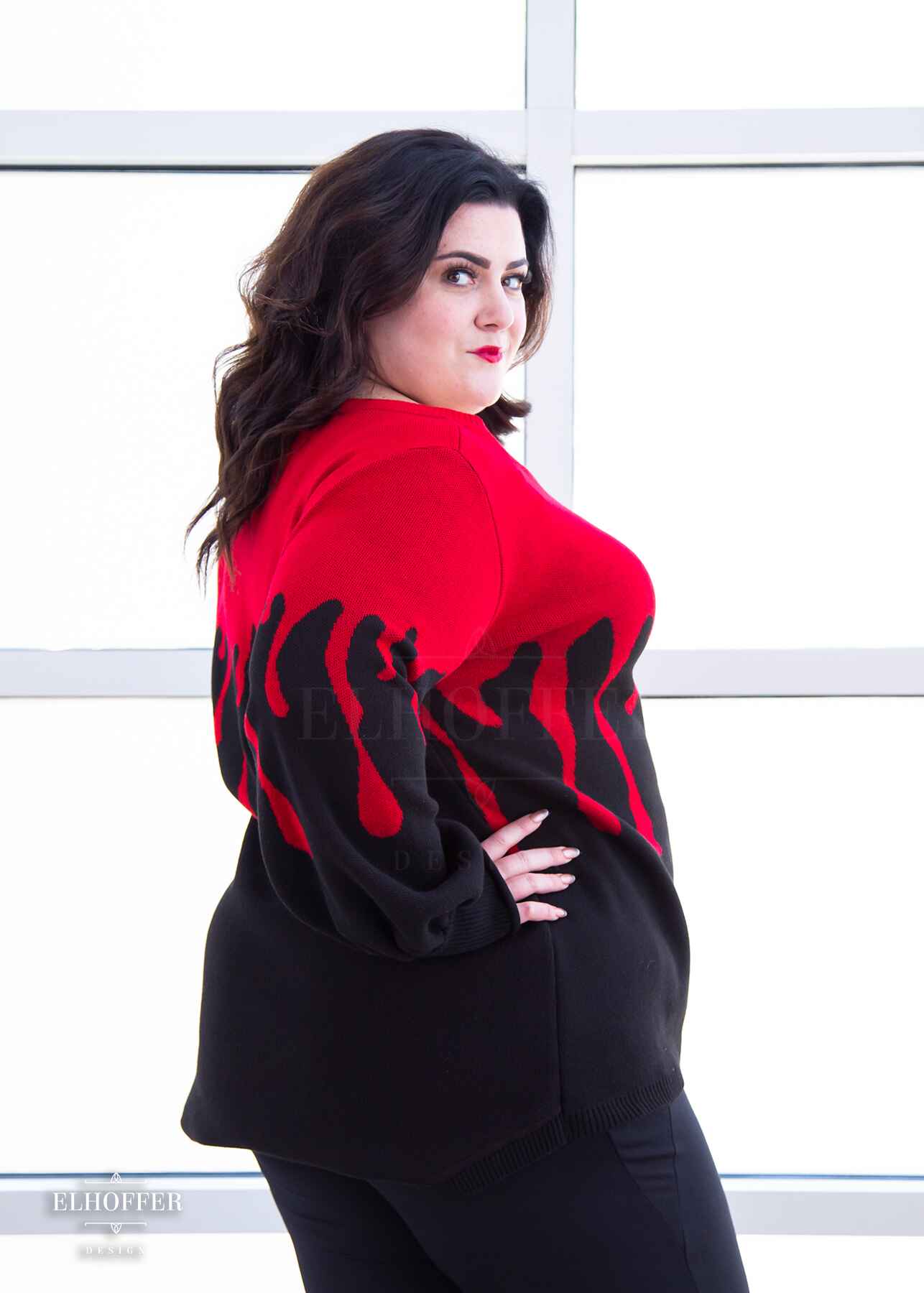 Stacy, a fair skinned XL model with shoulder length wavy dark brown hair, is wearing an oversize sweater with a bright red blood drip design that looks like it's oozing down onto a black sweater that has long billowing sleeves with thumbholes.
