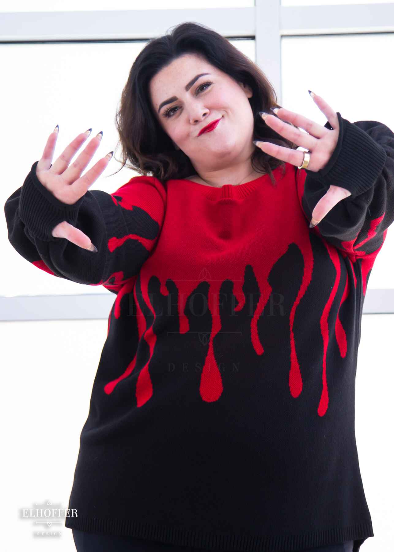 Stacy, a fair skinned XL model with shoulder length wavy dark brown hair, is smirking while holding her hands towards the camera to show off the thumbholes of the oversize sweater she is wearing that has a bright red blood drip design that looks like it's oozing down onto a black sweater with long billowing sleeves