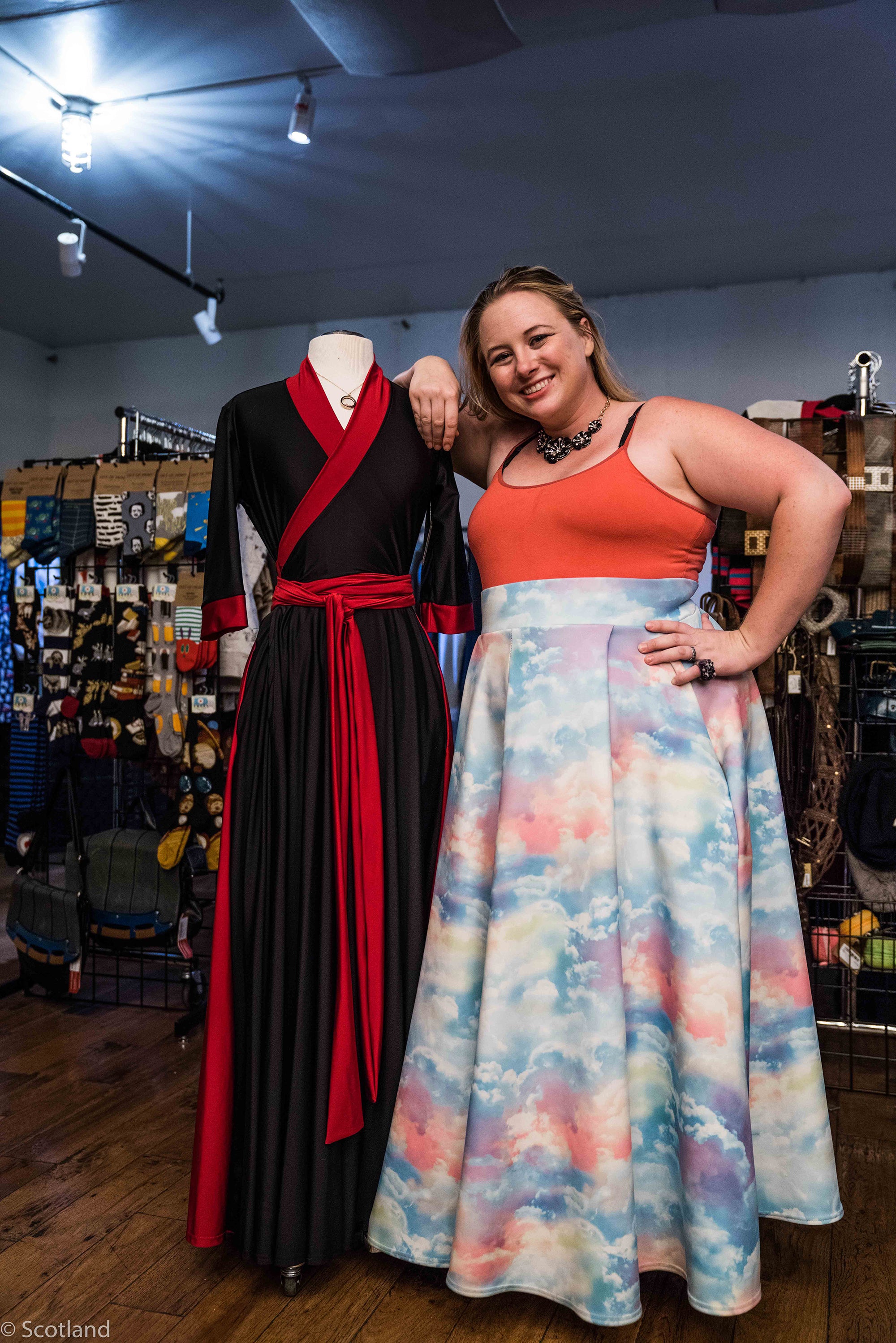 Catherine, a mid to plus sized white woman with blond hair, is wearing a coral tank and pastel rainbow cloud maxi skirt and is leaning against a mannequin wearing a black and red maxi wrap dress while standing in a boutique.