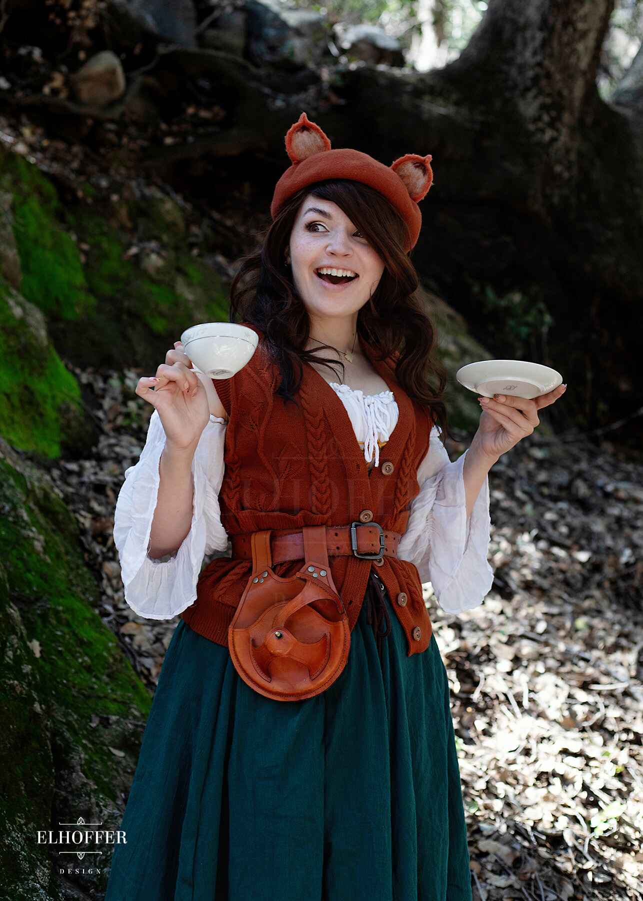 Kiri, a fair skinned S model with long dark brown wavy hair, is in the woods smiling while wearing a pumpkin orange button up knit vest with a leafy vine and cable knit pattern, light brown buttons, and front pockets.