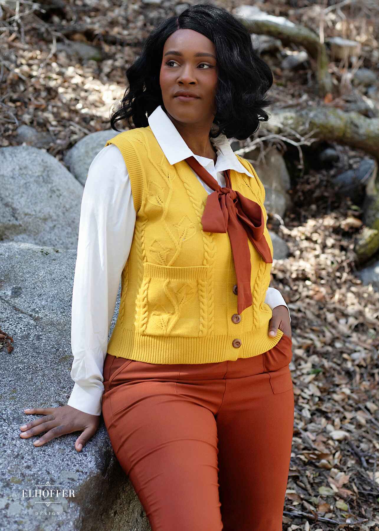 Lynsi, a medium brown skinned M model with shoulder length black hair, is in the woods wearing a golden yellow button up knit vest with a leafy vine and cable knit pattern, light brown buttons, and front pockets.
