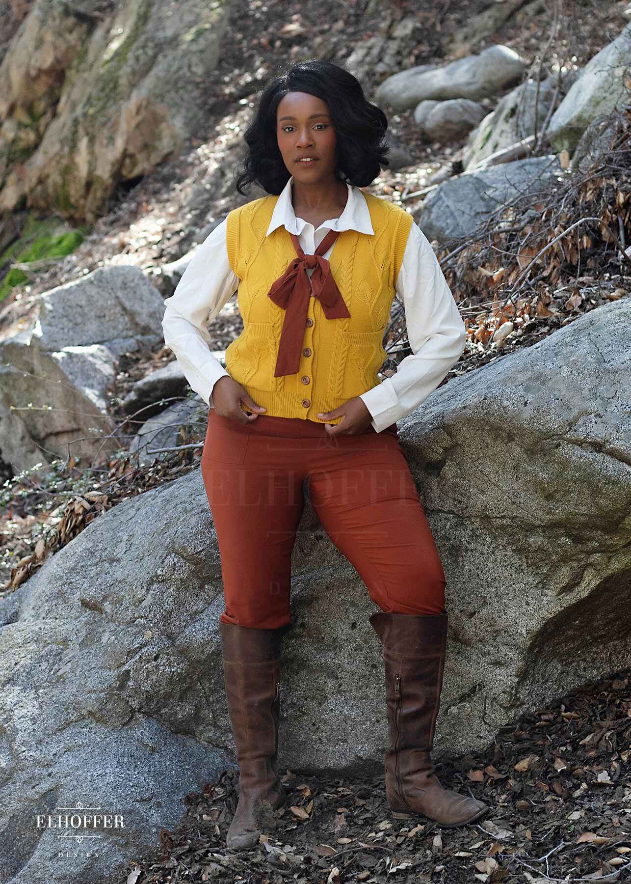 Lynsi, a medium brown skinned M model with shoulder length black hair, is in the woods wearing a golden yellow button up knit vest with a leafy vine and cable knit pattern, light brown buttons, and front pockets.