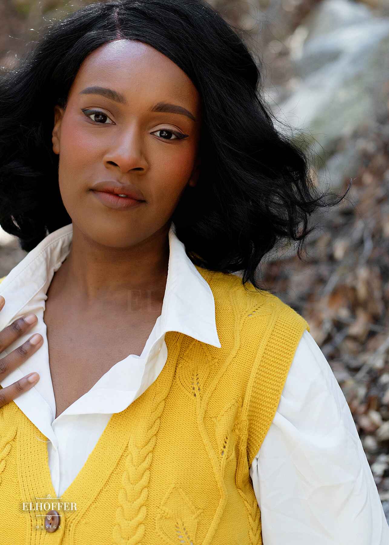 A closeup of Lynsi, a medium brown skinned M model with shoulder length black hair, wearing a golden yellow button up knit vest with a leafy vine and cable knit pattern, light brown buttons, and front pockets.