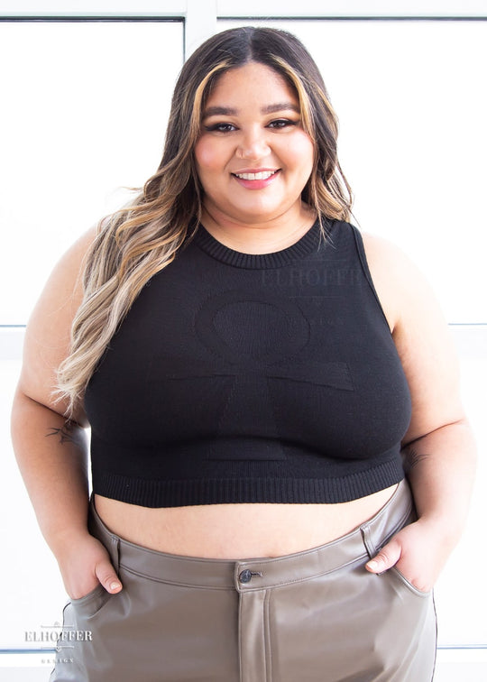 Cori, a sun kissed skinned 2xl model with long balayage hair, is smiling while wearing a sleeveless knit crop top with a large subtle ankh design on the front.