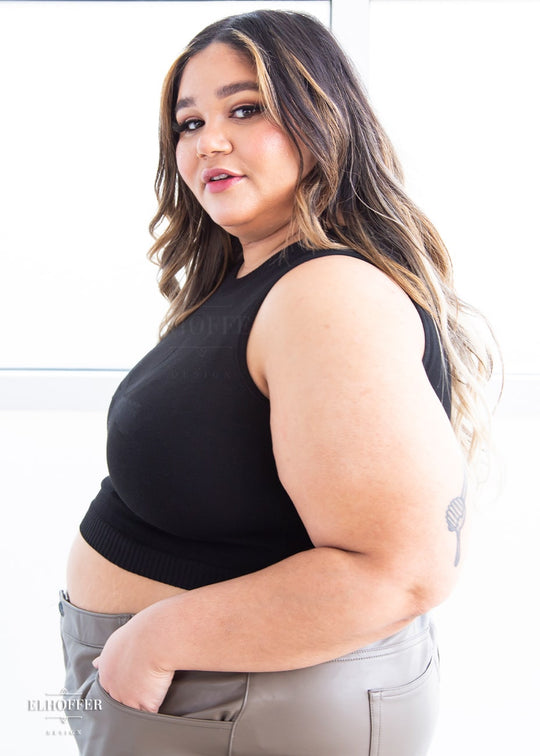 A side view of Cori, a sun kissed skinned 2xl model with long balayage hair, wearing a sleeveless knit crop top with a large subtle ankh design on the front.