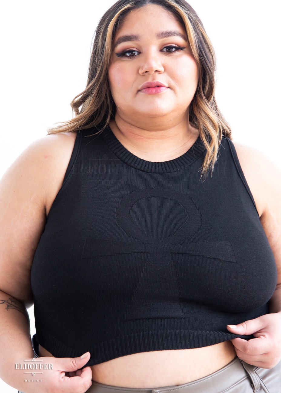 Cori, a sun kissed skinned 2xl model with long balayage hair, is wearing a sleeveless knit crop top with a large subtle ankh design on the front.