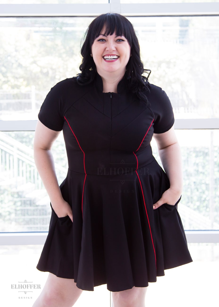 Bernadette, a fair skinned size large model with black hair and bangs, is wearing short raglan sleeved dress with a high neck, invisible front zipper, and princess seams on the front and back as well as pockets. The dress is black with red piping details.