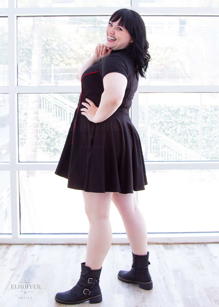 Bernadette, a fair skinned size large model with black hair and bangs, is wearing short raglan sleeved dress with a high neck, invisible front zipper, and princess seams on the front and back as well as pockets. The dress is black with red piping details.