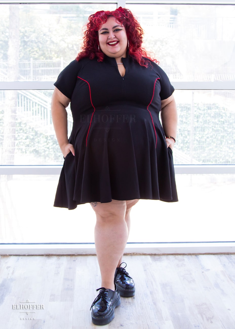 Victoria, an olive skinned size 4XL model with bright red curly hair, is wearing short raglan sleeved dress with a high neck, invisible front zipper, and princess seams on the front and back as well as pockets. The dress is black with red piping details.