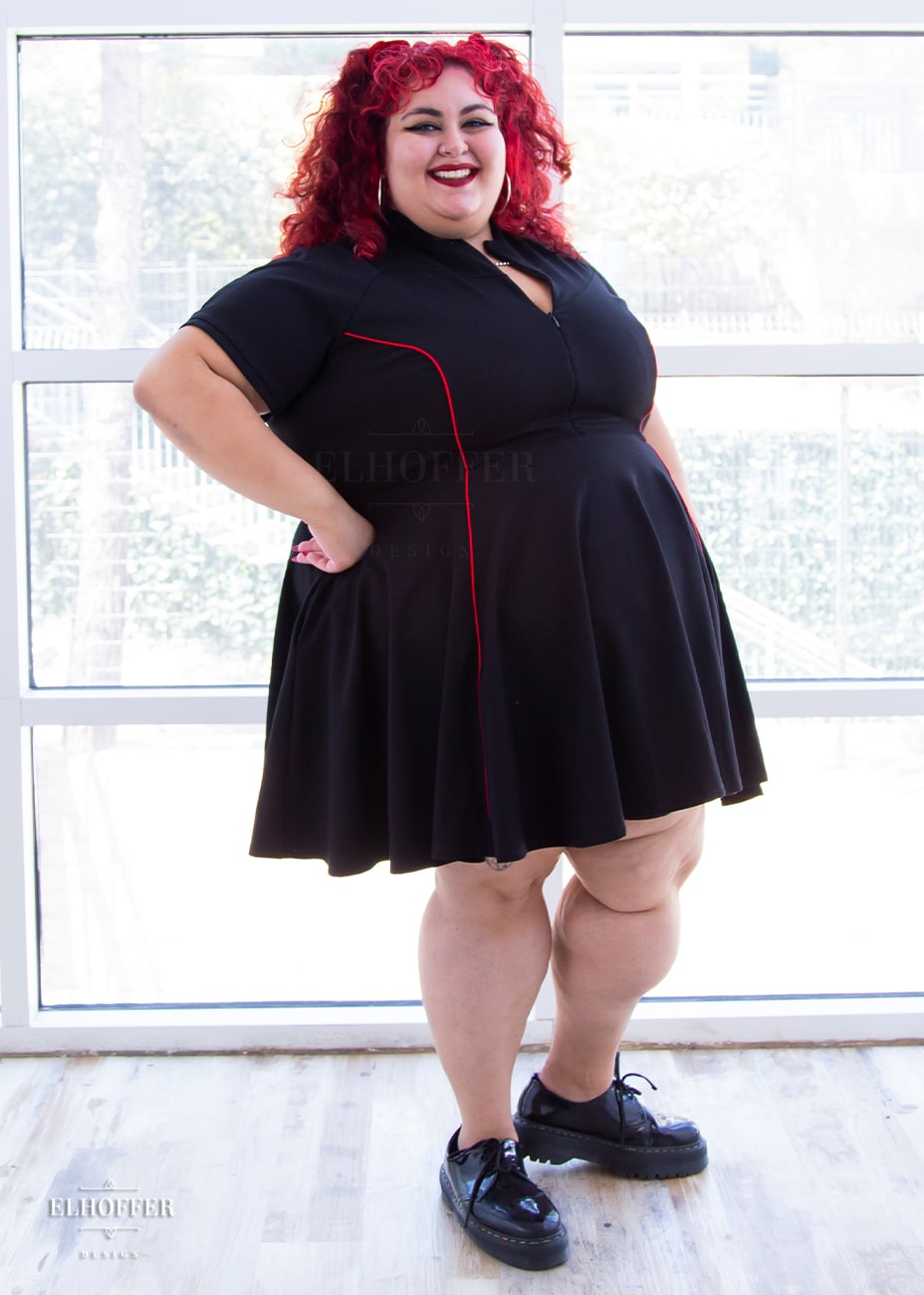 Victoria, an olive skinned size 4XL model with bright red curly hair, is wearing short raglan sleeved dress with a high neck, invisible front zipper, and princess seams on the front and back as well as pockets. The dress is black with red piping details.