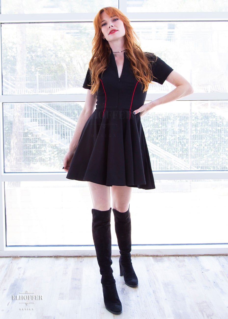 Chloe, a fair skinned size XS model with red hair and bangs, is wearing short raglan sleeved dress with a high neck, invisible front zipper, and princess seams on the front and back as well as pockets. The dress is black with red piping details.