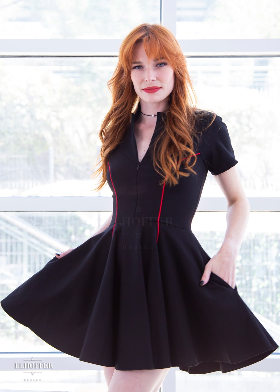Chloe, a fair skinned size XS model with red hair and bangs, is wearing short raglan sleeved dress with a high neck, invisible front zipper, and princess seams on the front and back as well as pockets. The dress is black with red piping details.