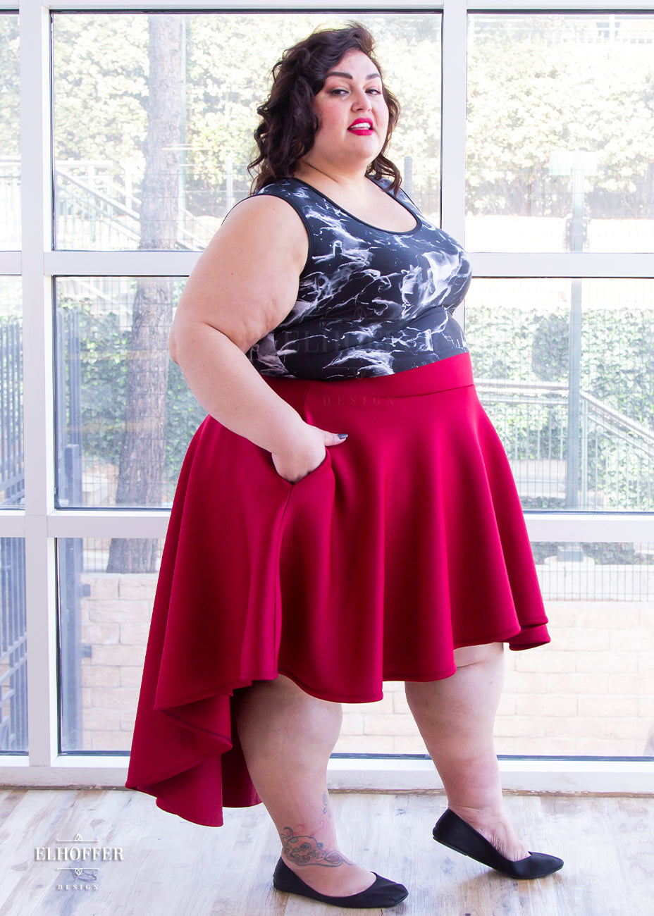 Kristen, a size 3XL olive skinned model with short dark brown hair, is wearing a burgundy commander skirt and a smoke print black and white tank. The skirt is a high waisted hi-low skirt that stands away from the body due to the double scuba fabric with encased elastic waistband with matching lining.