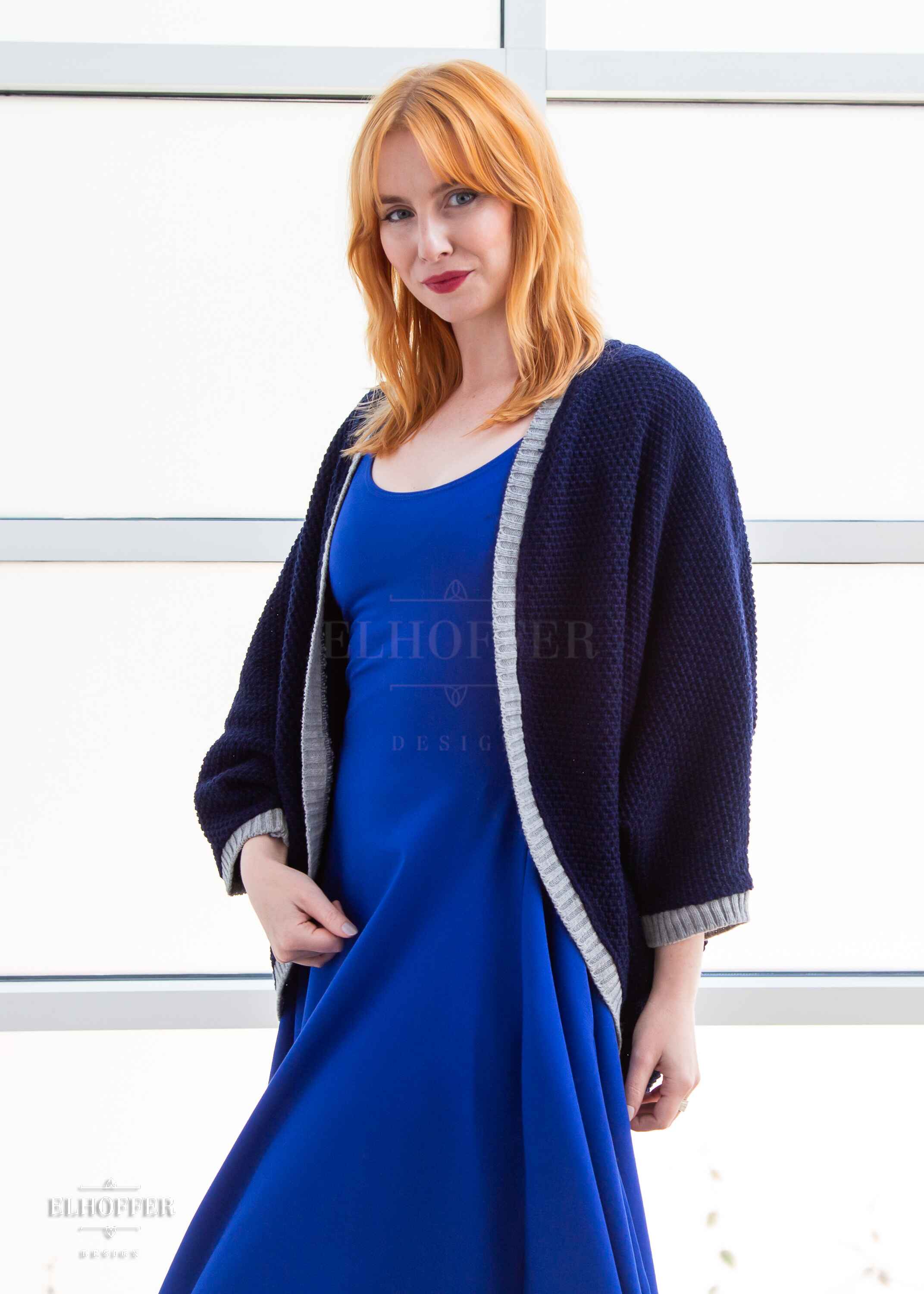Harley, a fair skinned S model with shoulder length strawberry blonde hair, is wearing a dark blue loose knit dolman with light grey ribbing around edges and cuffs. The dolman features 3/4 length sleeves and a magical script design (circle with T crossing through the circle) on the back. She paired the dolman with a cobalt blue knee length dress.