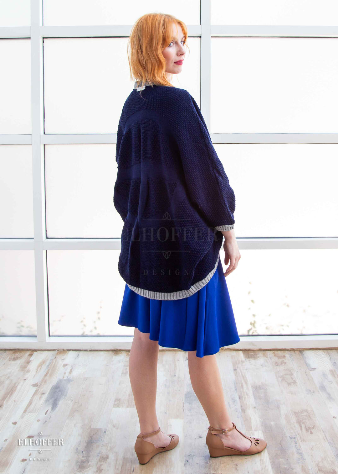 Harley, a fair skinned S model with shoulder length strawberry blonde hair, is wearing a dark blue loose knit dolman with light grey ribbing around edges and cuffs. The dolman features 3/4 length sleeves and a magical script design (circle with T crossing through the circle) on the back. She paired the dolman with a cobalt blue knee length dress.