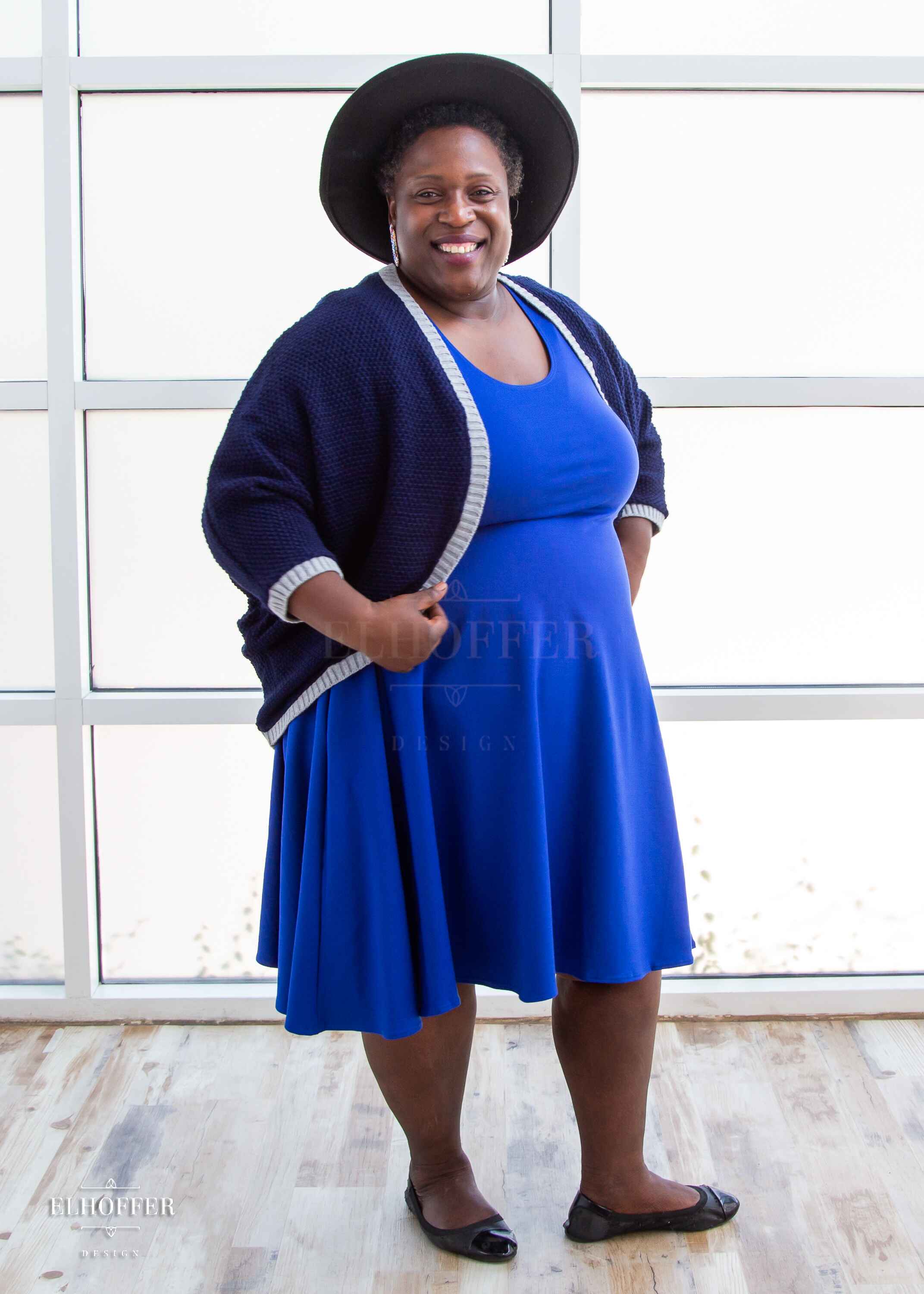 Adalgize, a medium dark skinned 4xl model with short super curly black hair, is smiling while wearing a dark blue loose knit dolman with light grey ribbing around edges and cuffs.  The dolman features 3/4 length sleeves and a magical script design (circle with a T crossing through the circle) on the back.  She paired the dolman with a cobalt blue knee length dress.