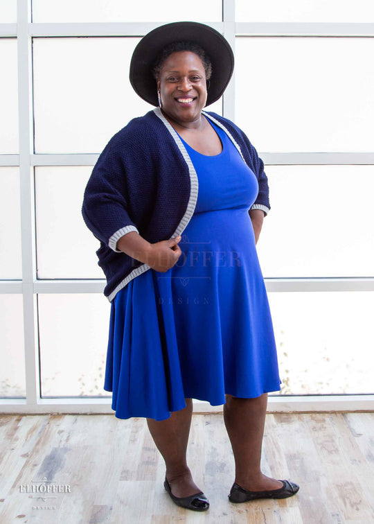 Adalgize, a medium dark skinned 4xl model with short super curly black hair, is smiling while wearing a dark blue loose knit dolman with light grey ribbing around edges and cuffs.  The dolman features 3/4 length sleeves and a magical script design (circle with a T crossing through the circle) on the back.  She paired the dolman with a cobalt blue knee length dress.