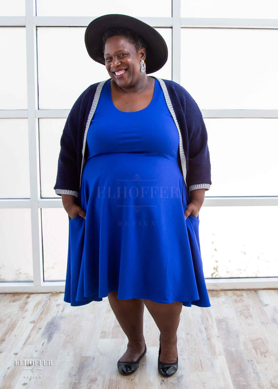 Adalgize, a medium dark skinned 4xl model with short super curly black hair, is smiling while wearing a dark blue loose knit dolman with light grey ribbing around edges and cuffs.  The dolman features 3/4 length sleeves and a magical script design (circle with a T crossing through the circle) on the back.  She paired the dolman with a cobalt blue knee length dress.