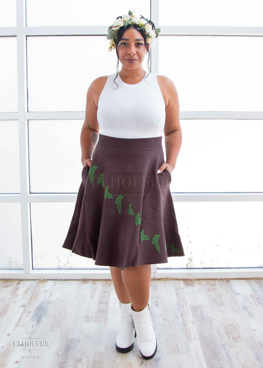 Janae, a light brown skinned L model with dark brown hair in an updo, is wearing a brown knit skirt that hits just passed the knee and has a cascading green leaf design and a white knit crop top with a butterly wing texture...