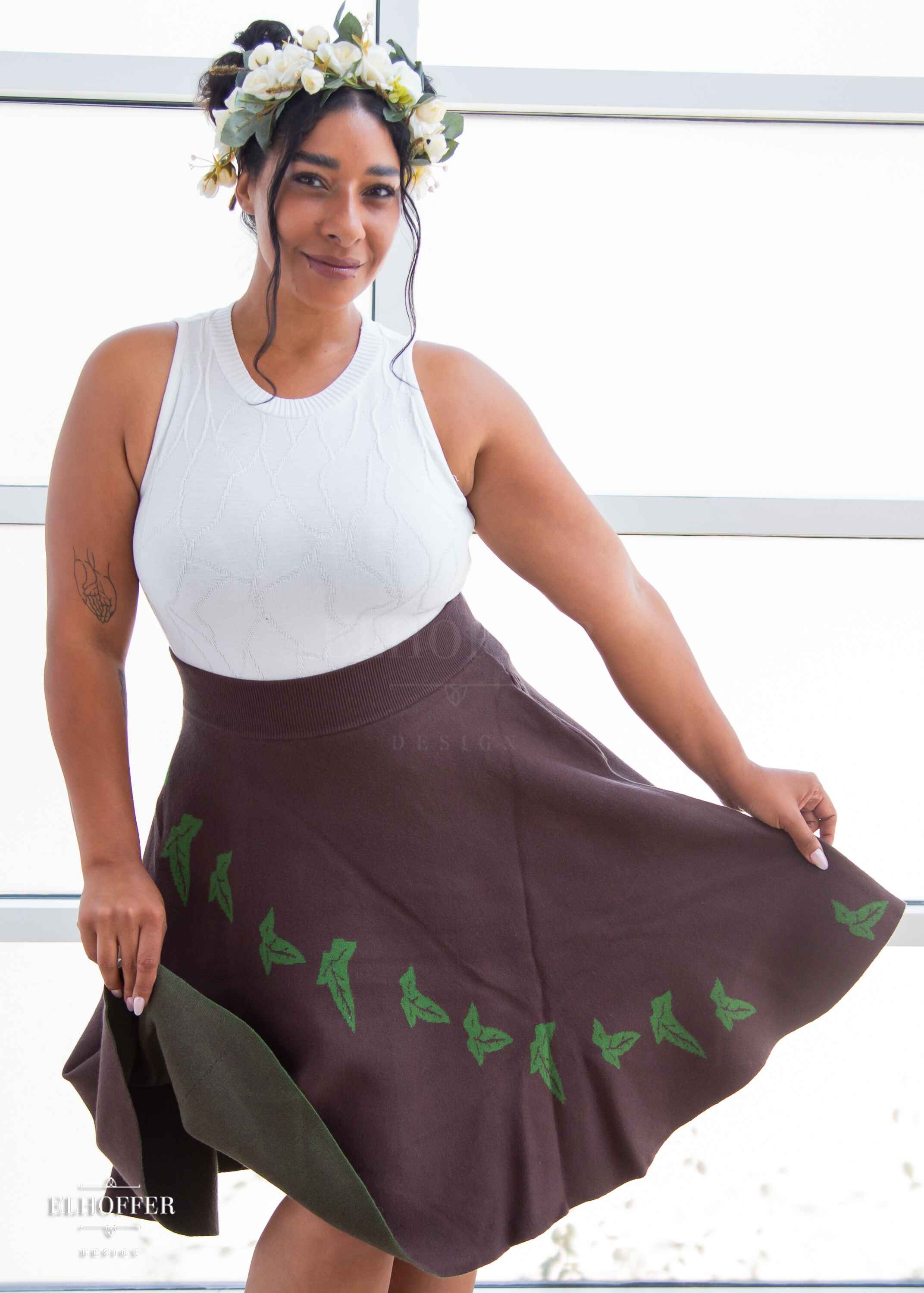 Janae, a light brown skinned L model with dark brown hair in an updo, is wearing a brown knit skirt that hits just passed the knee and has a cascading green leaf design and a white knit crop top with a butterly wing texture...