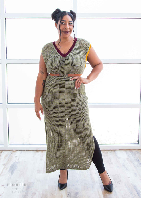 Janae, a light brown skinned L model with dark brown hair in space buns, is wearing a long reversible sleeveless loose knit cover up with side slits.  The main body is an olive green with one side plain knit with a v neck and the other side having a subtle skull design with a scoop neck, the neckline is trimmed in crimson red, and the armholes are trimmed in a yellow.