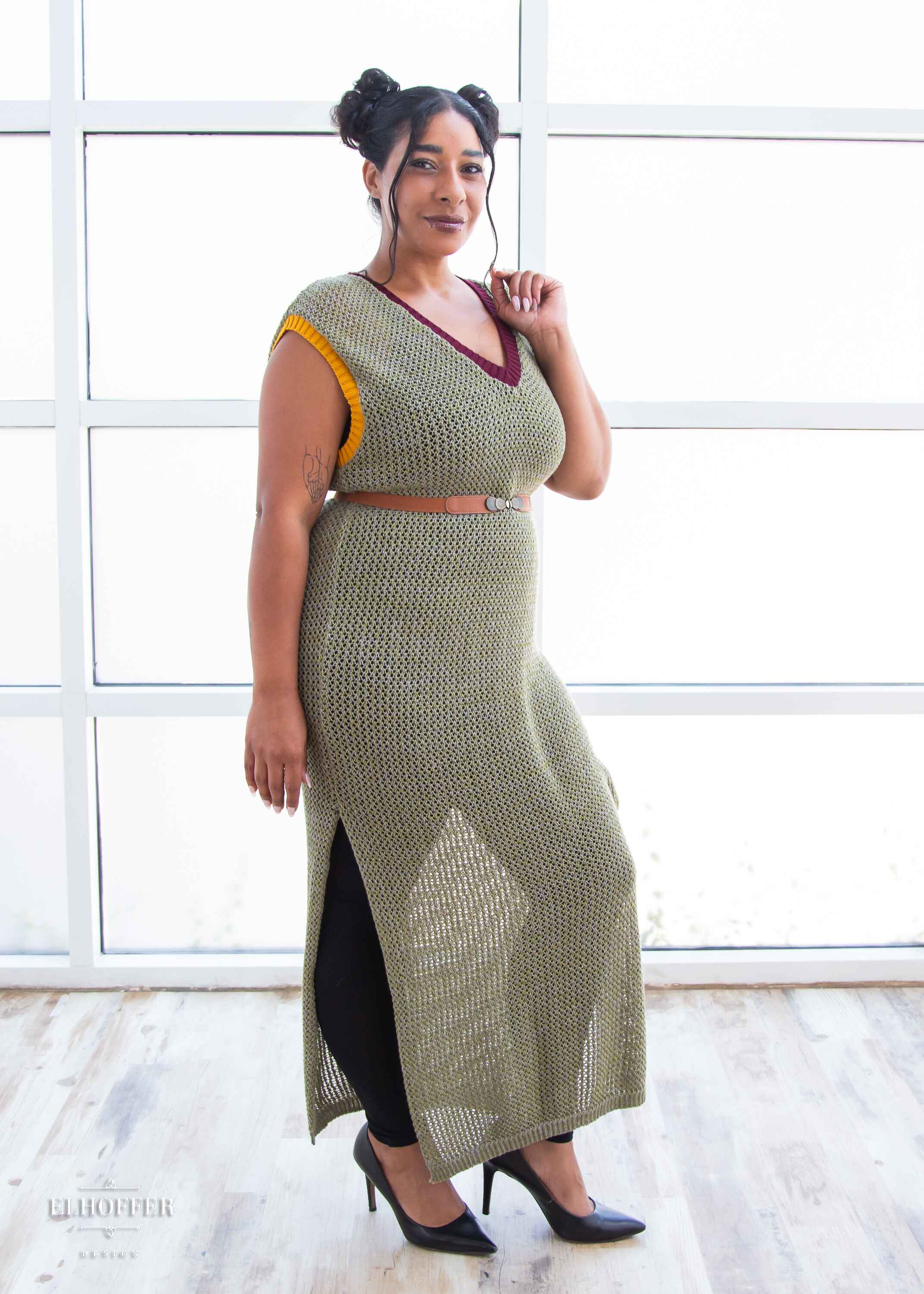 Janae, a light brown skinned L model with dark brown hair in space buns, is wearing a long reversible sleeveless loose knit cover up with side slits.  The main body is an olive green with one side plain knit with a v neck and the other side having a subtle skull design with a scoop neck, the neckline is trimmed in crimson red, and the armholes are trimmed in a yellow.