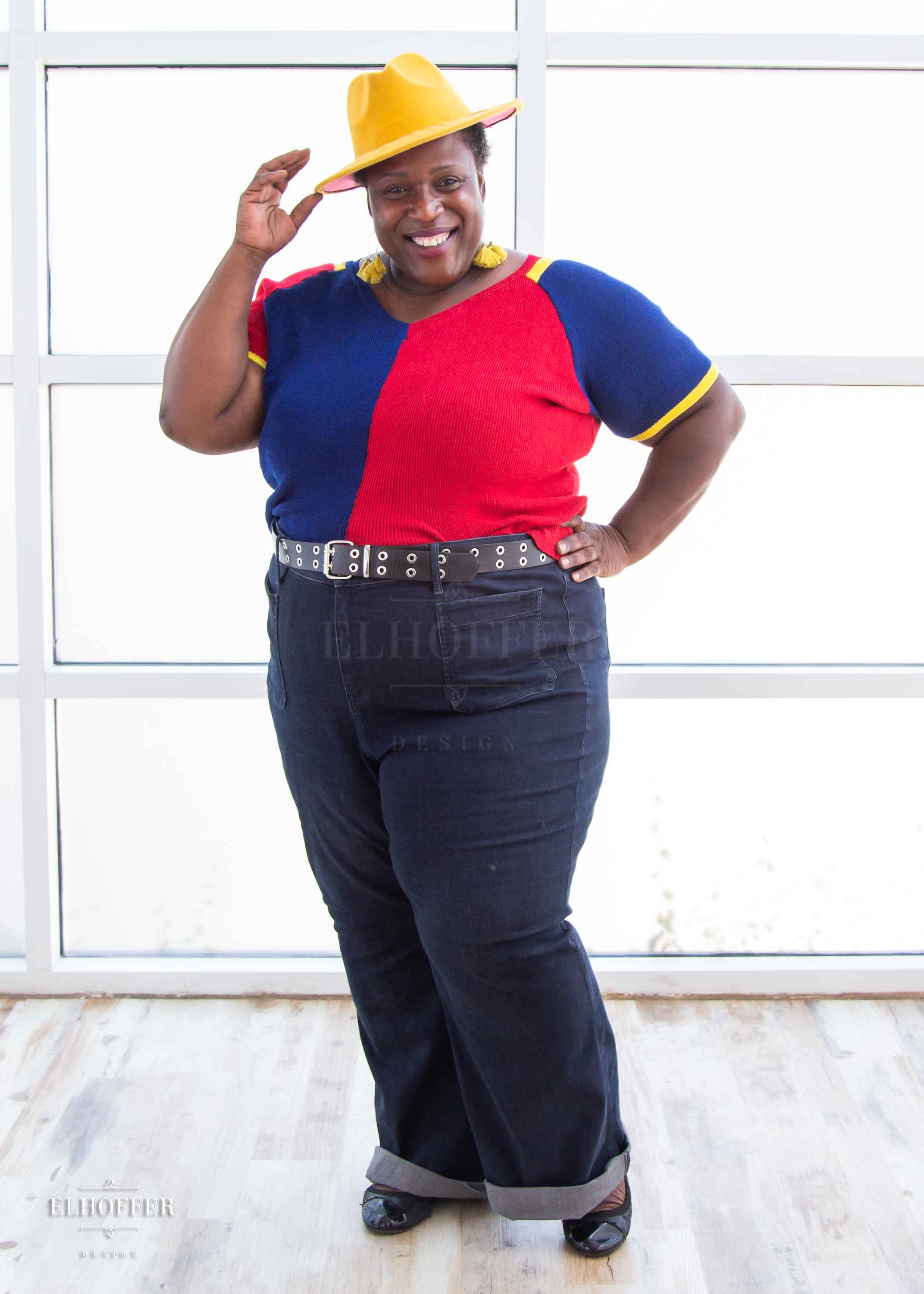Adalgiza, a medium dark skinned 4xl model with short black super curly hair, is smiling while wearing a short sleeve knit top with alternating blue and red colors. There is yellow detailing along the top of the shoulder and around the cuff of the sleeve.