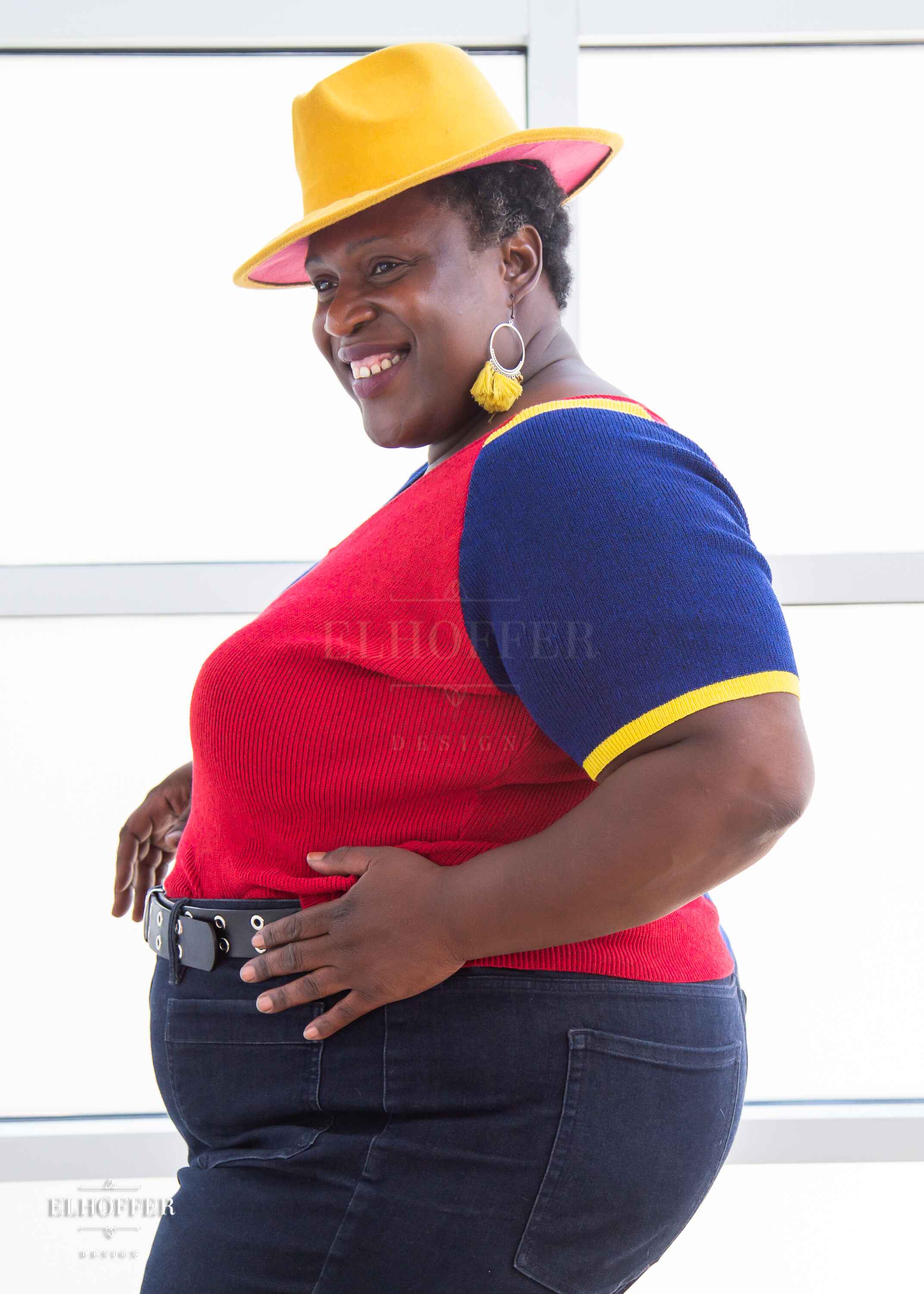 Adalgiza, a medium dark skinned 4xl model with short black super curly hair, is smiling while wearing a short sleeve knit top with alternating blue and red colors. There is yellow detailing along the top of the shoulder and around the cuff of the sleeve.