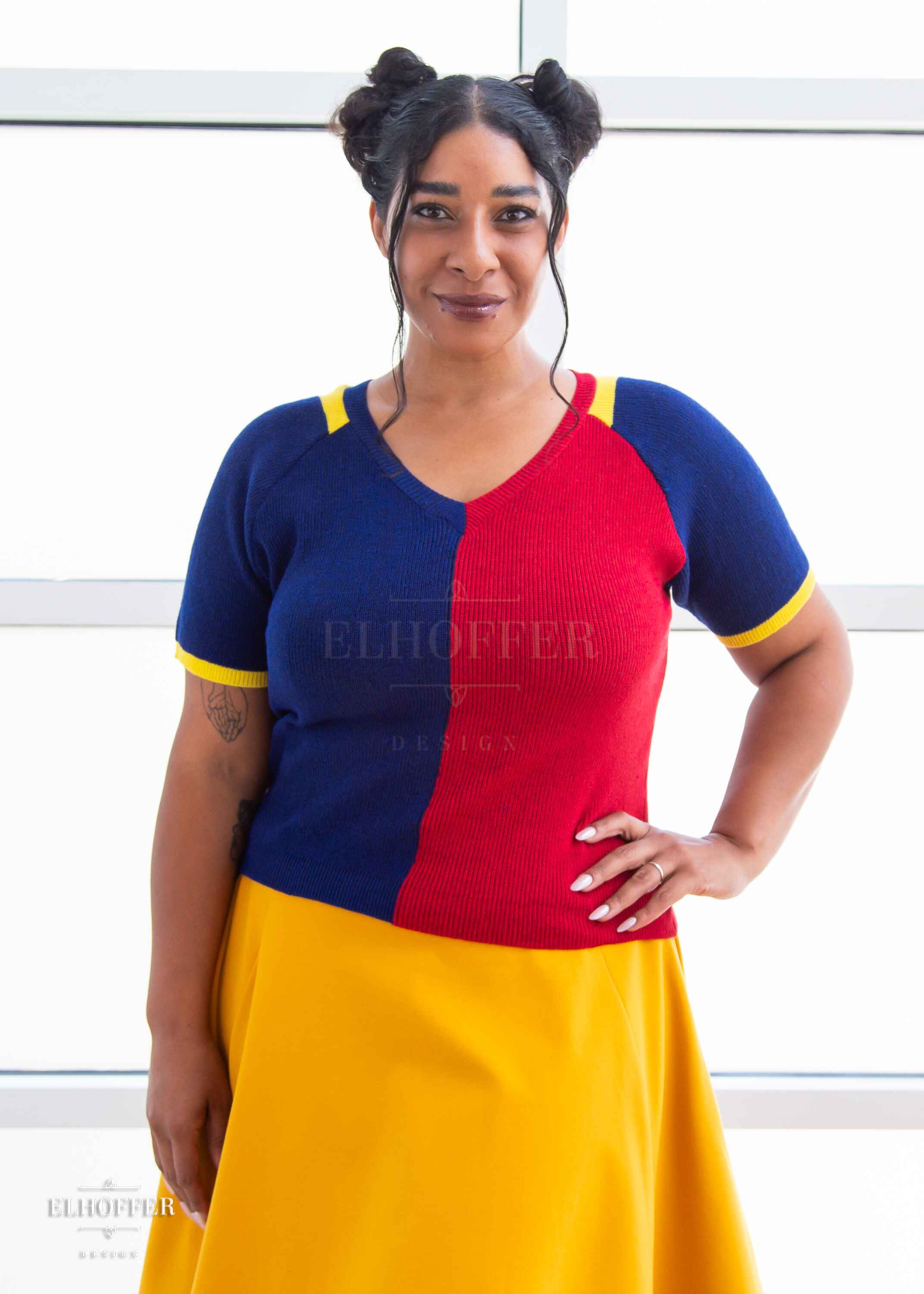 Janae, a light brown skinned L model with dark brown hair in space buns, is wearing a short sleeve knit top with alternating blue and red colors. There is yellow detailing along the top of the shoulder and around the cuff of the sleeve. She paired the knit top with a golden yellow knee length high low skirt.