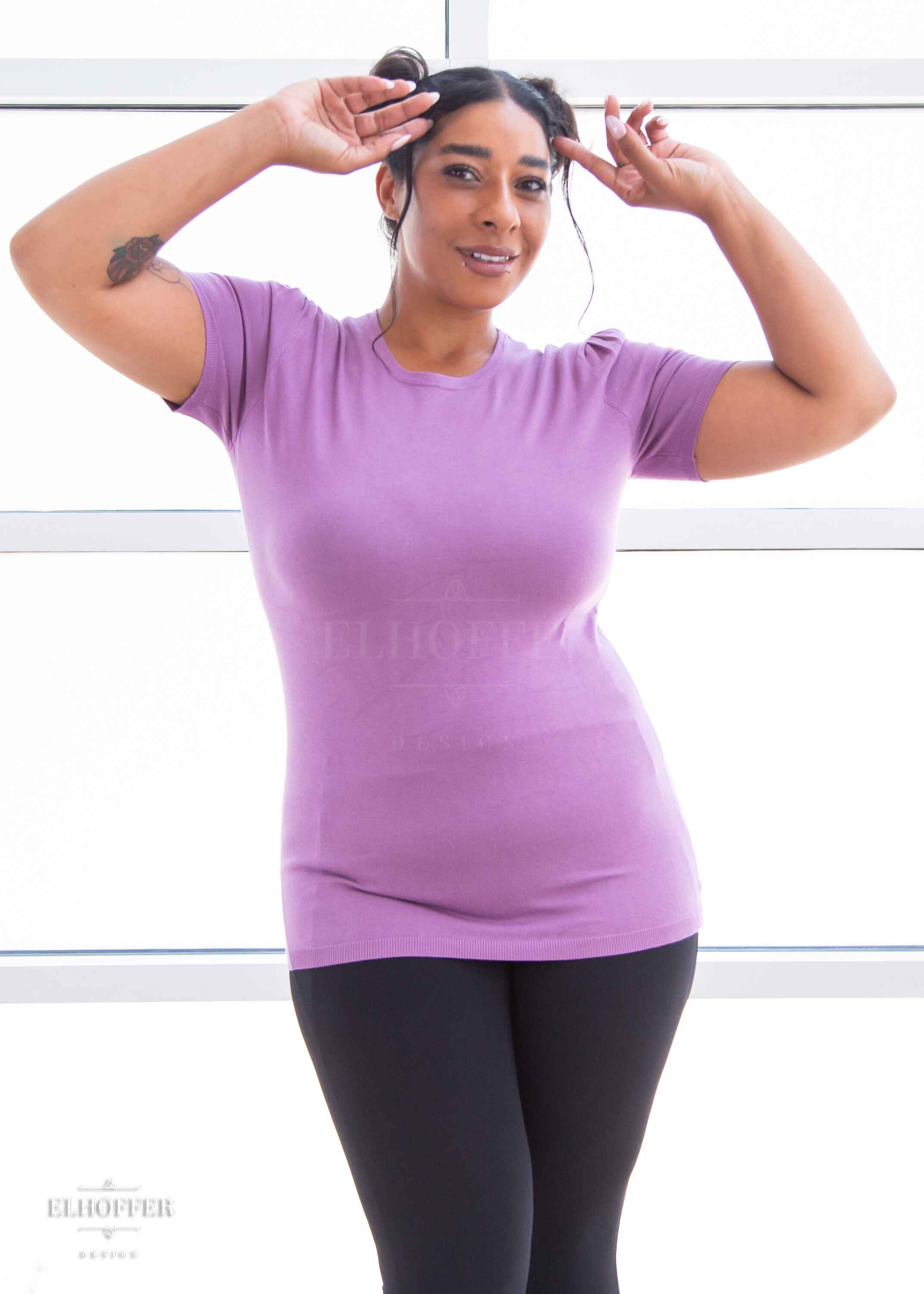 Janae, a light brown skinned L model with dark hair up in space buns, is wearing a short sleeve light weight soft purple knit top. The top hits about mid hip in length and the sleeves have pleated gathering at the shoulders.