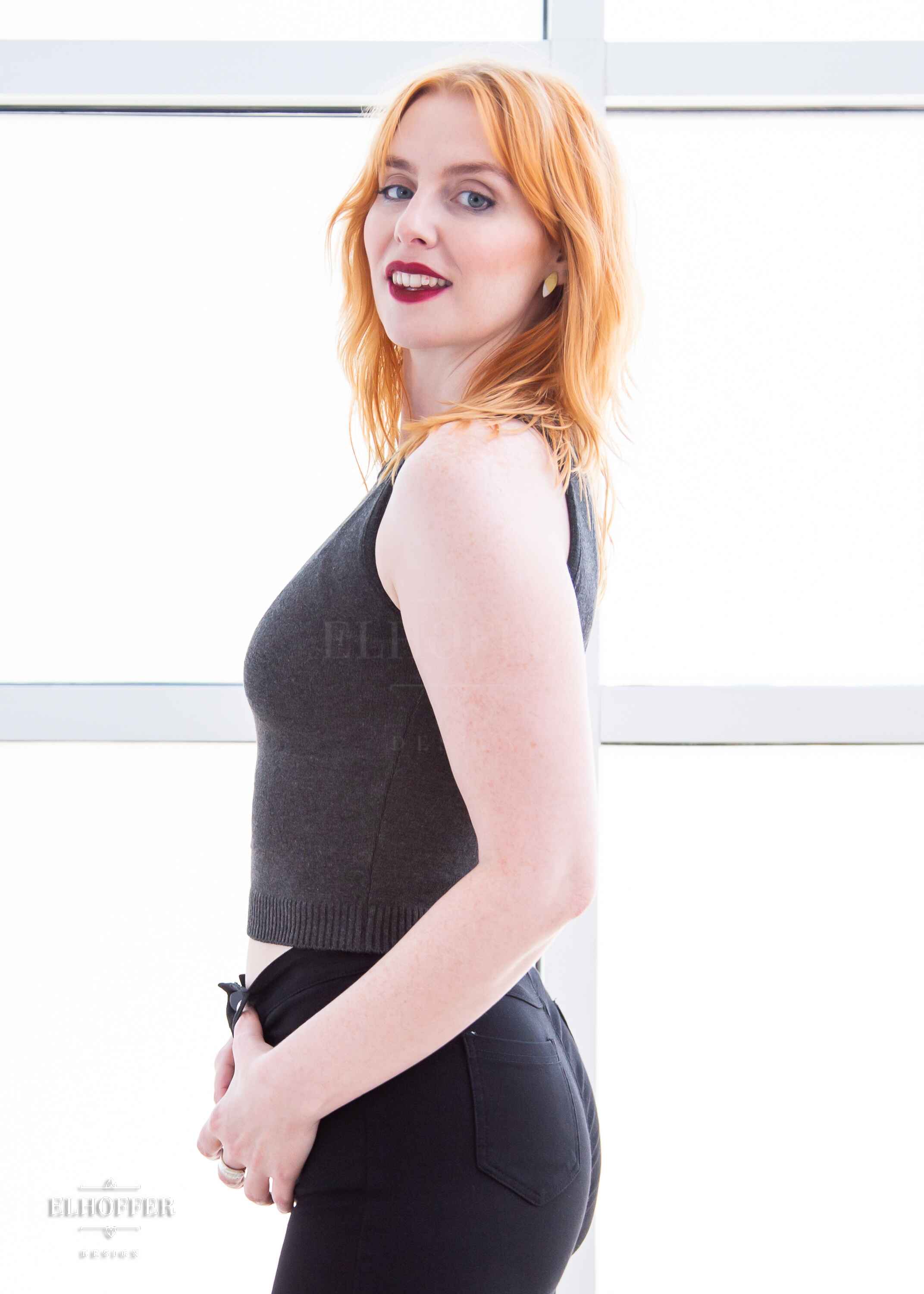 A side view of Harley, a fair skinned S model with shoulder length strawberry blonde hair, wearing a dark grey sleeveless knit crop top with a large subtle ankh design on the front.