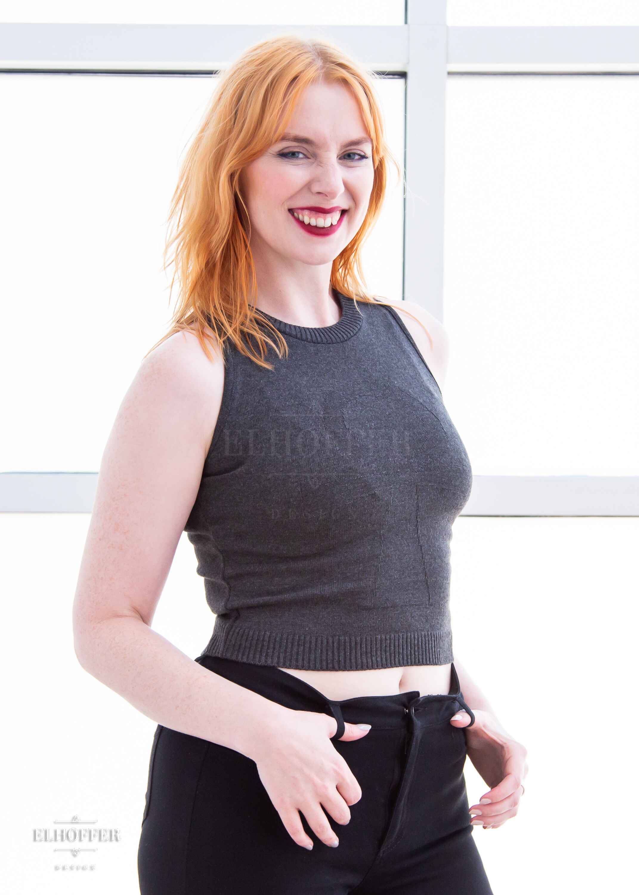 Harley, a fair skinned S model with shoulder length strawberry blonde hair, is smiling while wearing a dark grey sleeveless knit crop top with a large subtle ankh design on the front.