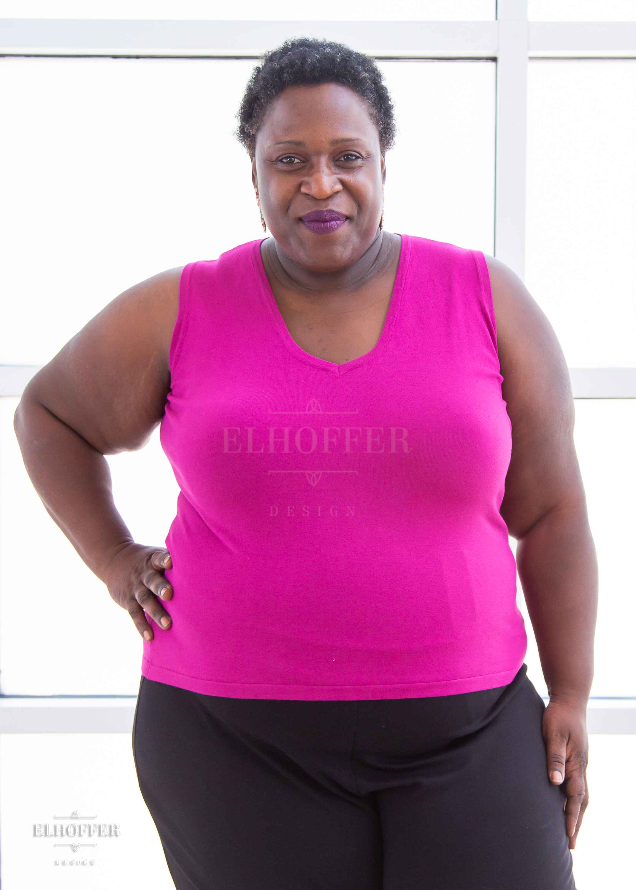 Adalgiza, a medium brown skinned 4xl model with short dark super curly hair, is smiling while wearing a bright pink lightweight knit v neck tank top. The tank top hits about mid hip in length.