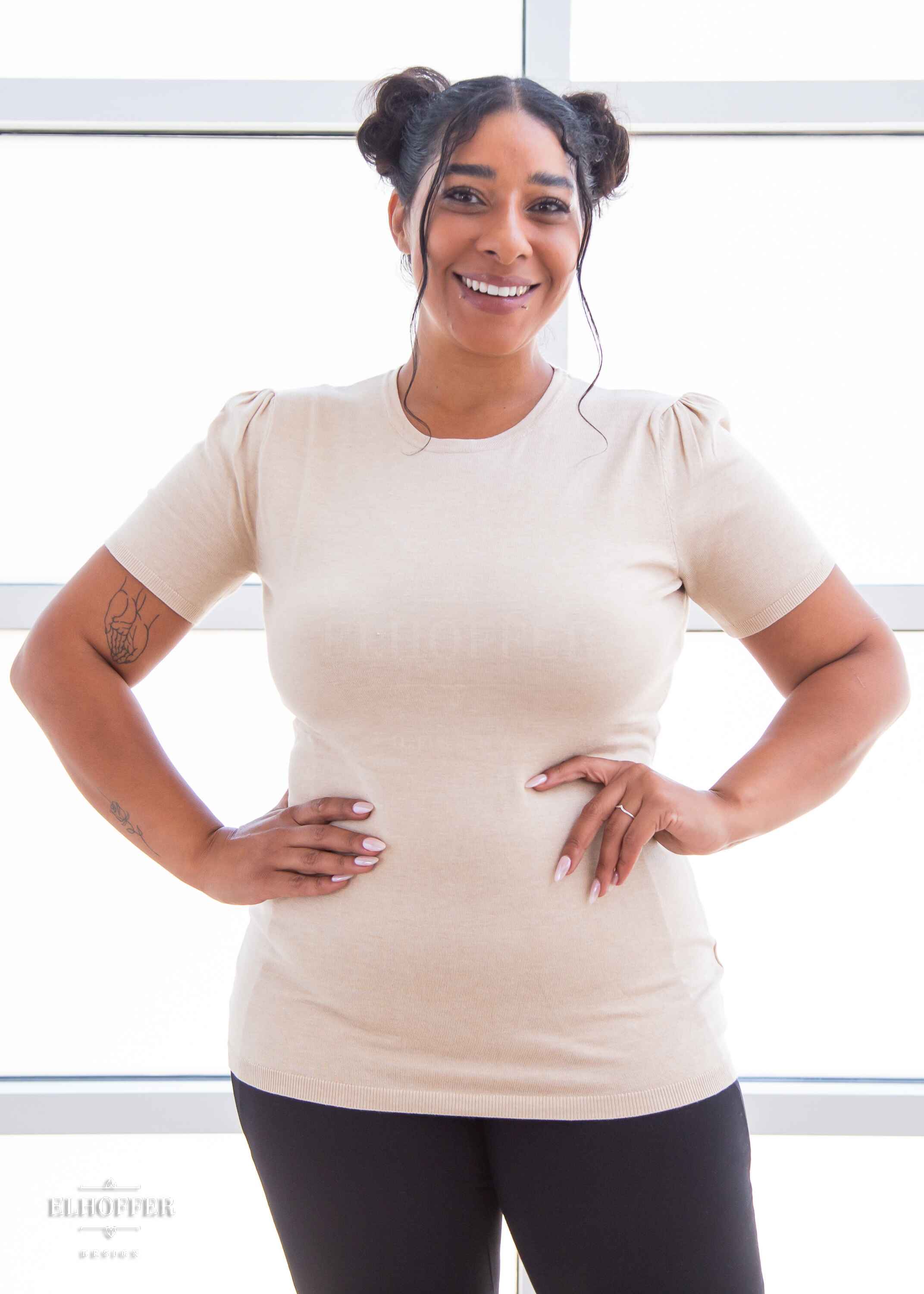 Janae, a light brown skinned L model with dark hair in space buns, is smiling while wearing a short sleeve light weight off white knit top. The top hits about mid hip in length and the sleeves have pleated gathering at the shoulders.