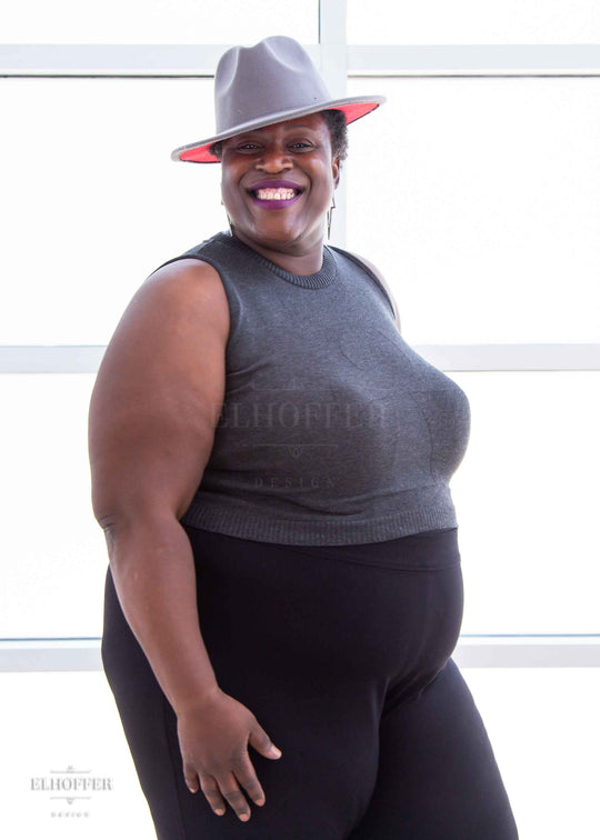 Adalgiza, a medium dark skinned 4xl model with short dark super curly hair, is smiling while wearing a dark grey sleeveless knit crop top with a large subtle ankh design on the front.