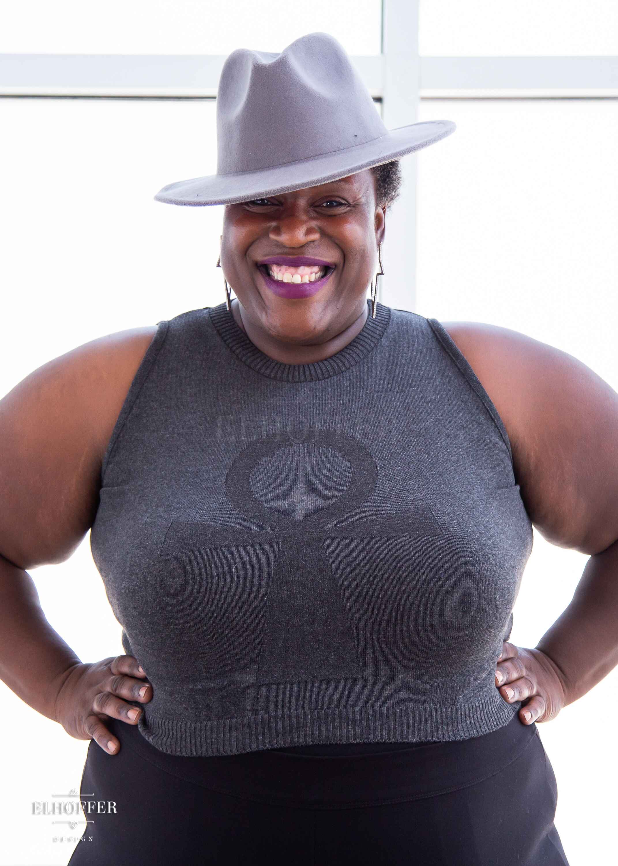 Adalgiza, a medium dark skinned 4xl model with short dark super curly hair, is smiling while wearing a dark grey sleeveless knit crop top with a large subtle ankh design on the front.