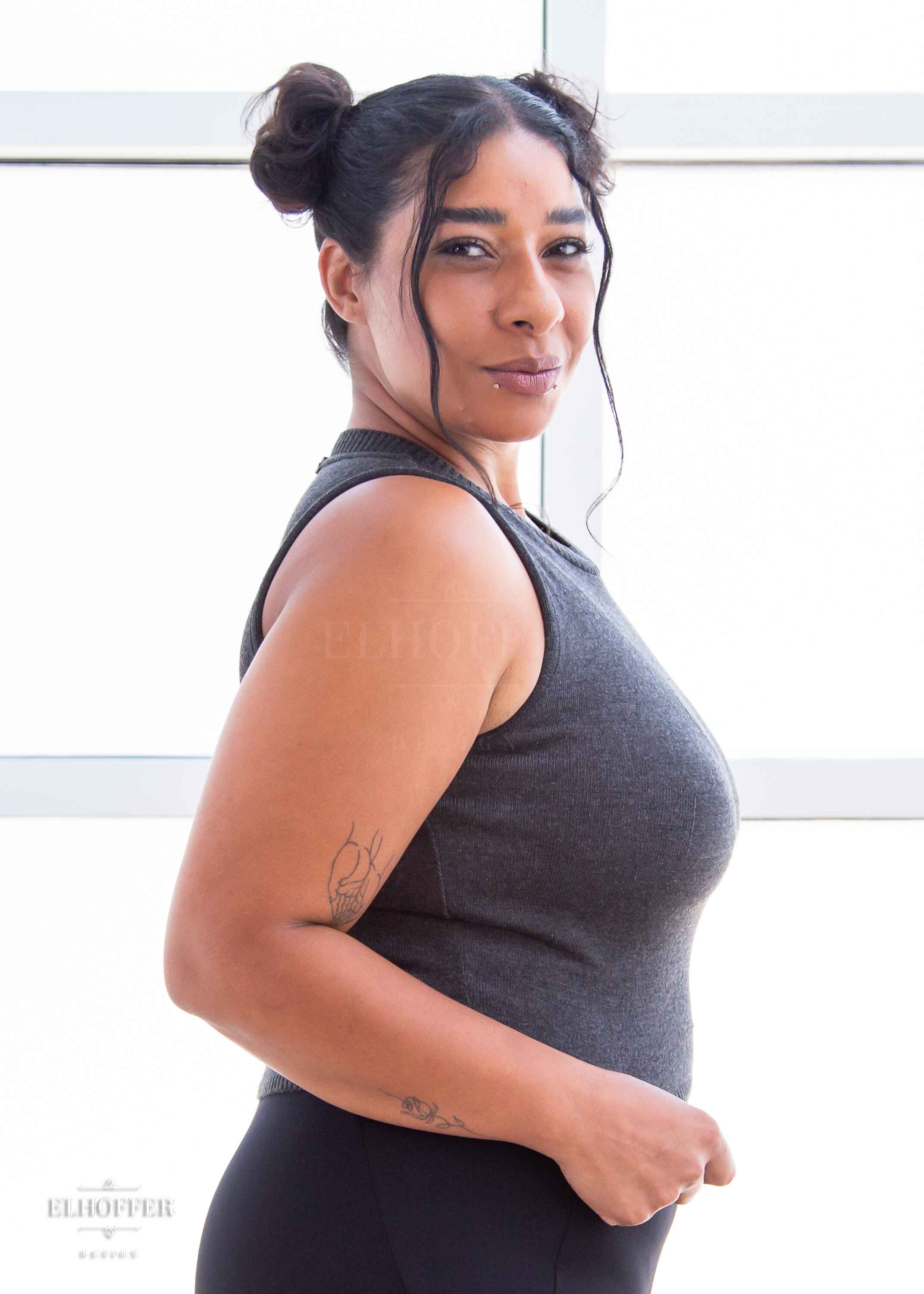 A side view of Janae, a light brown skinned L model with dark hair in space buns, wearing a dark grey sleeveless knit crop top with a large subtle ankh design on the front.