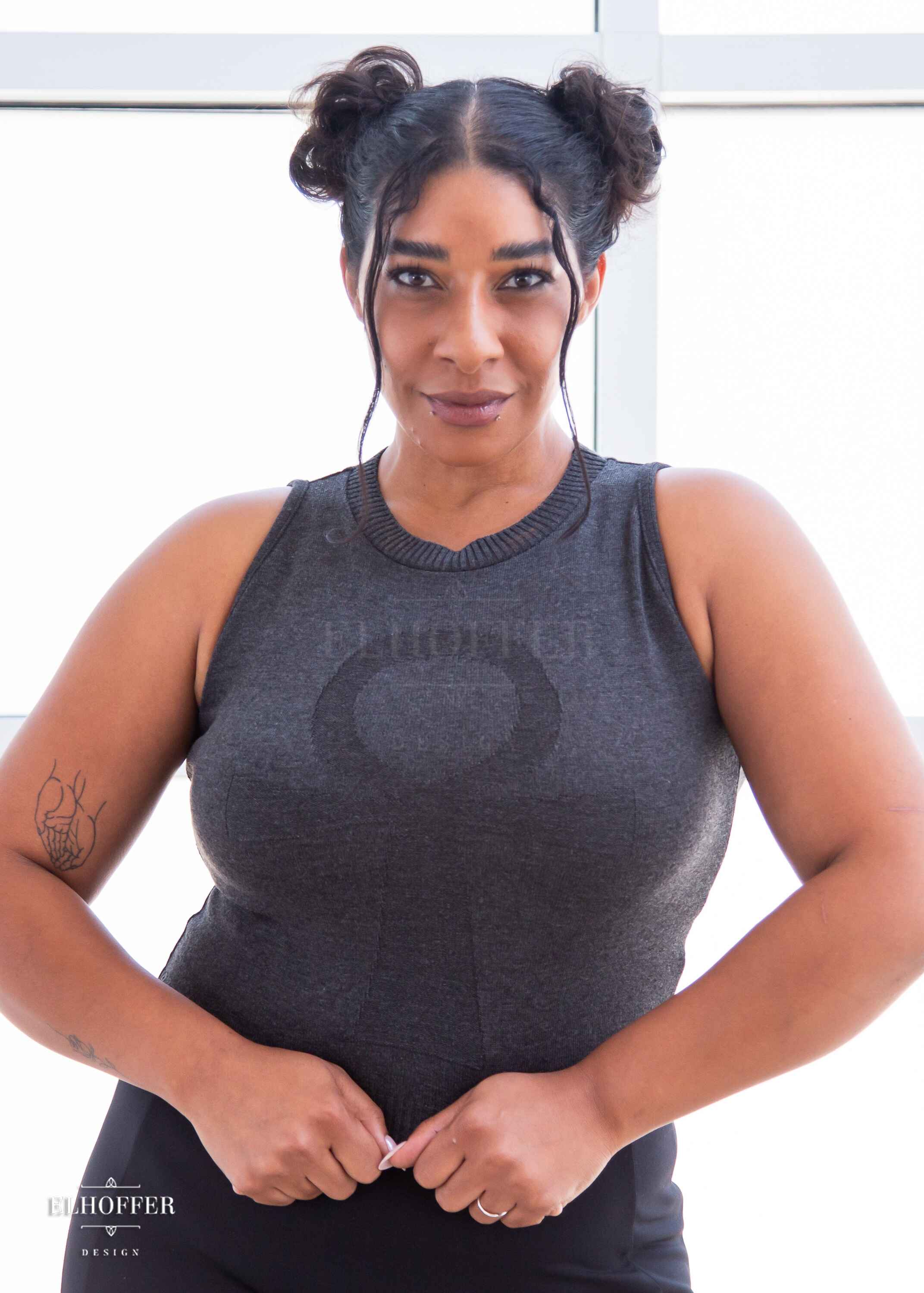 Janae, a light brown skinned L model with dark hair in space buns, is smiling while wearing a dark grey sleeveless knit crop top with a large subtle ankh design on the front.