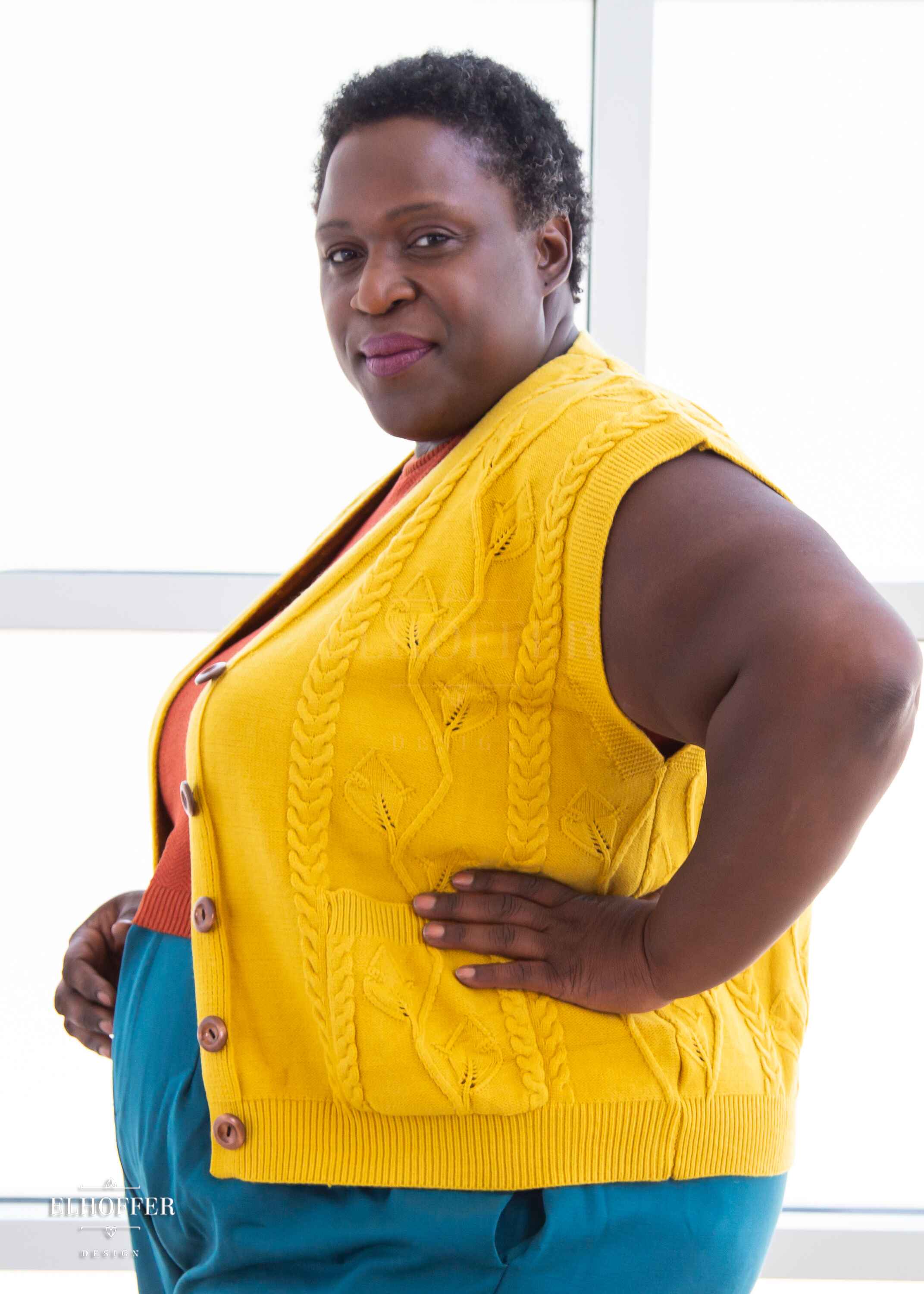 A side view of Adalgiza, a dark brown skinned 3xl model with short black tight curly hair, wearing a golden yellow button up knit vest with a leafy vine and cable knit pattern, light brown buttons, and front pockets. She paired the vest with a rust color knit crop top and peacock teal trousers.