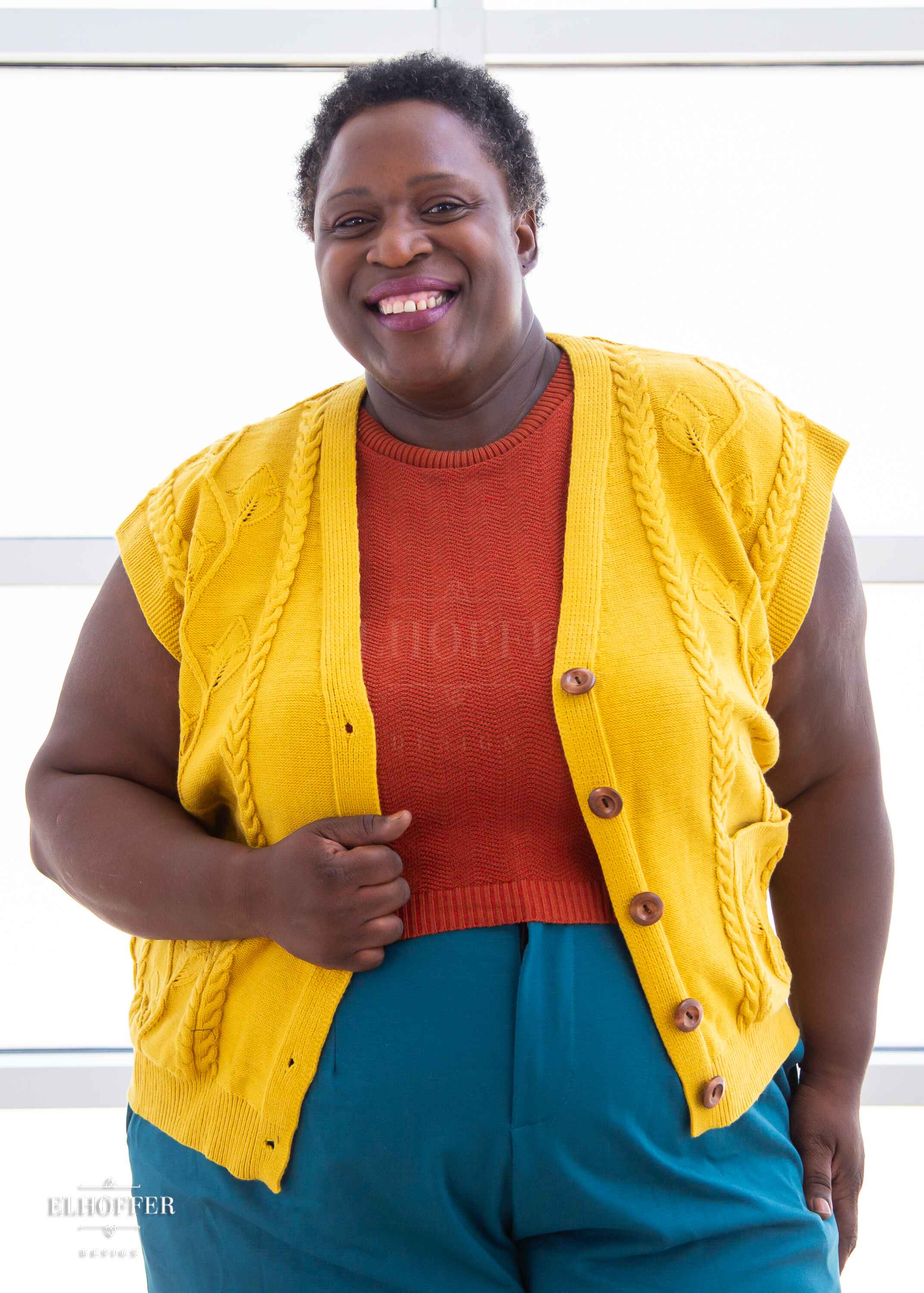 Adalgiza, a dark brown skinned 3xl model with short black tight curly hair, is smiling while wearing a golden yellow button up knit vest with a leafy vine and cable knit pattern, light brown buttons, and front pockets. She paired the vest with a rust color knit crop top and peacock teal trousers.