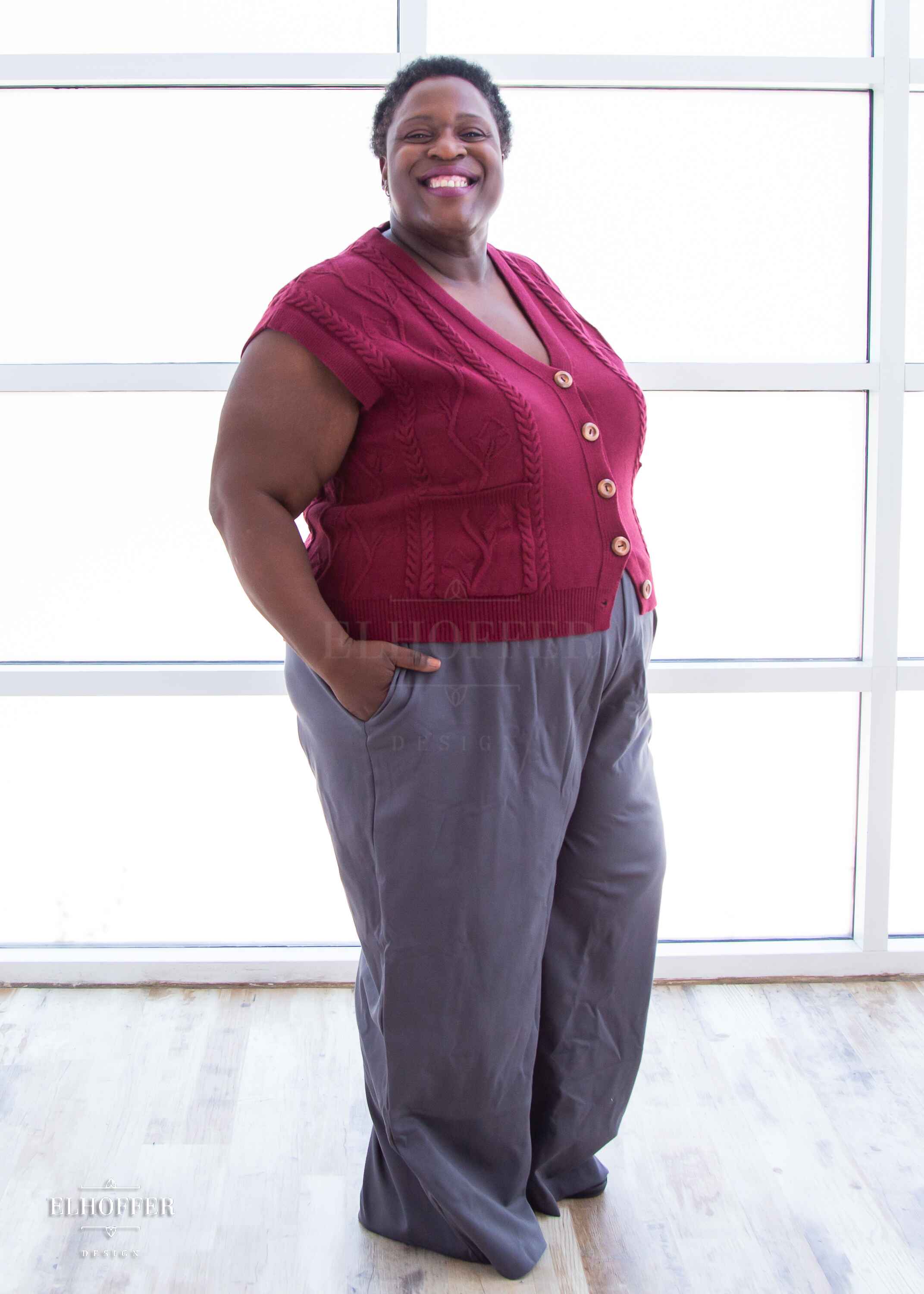 Adalgiza, a medium dark skinned 4xl model with short dark super curly hair, is smiling while wearing the L (3XL-4XL) sample of a cranberry red button up knit vest with a leafy vine and cable knit pattern, light brown buttons, and front pockets. She paired the knit vest with charcoal grey pleated trousers.
