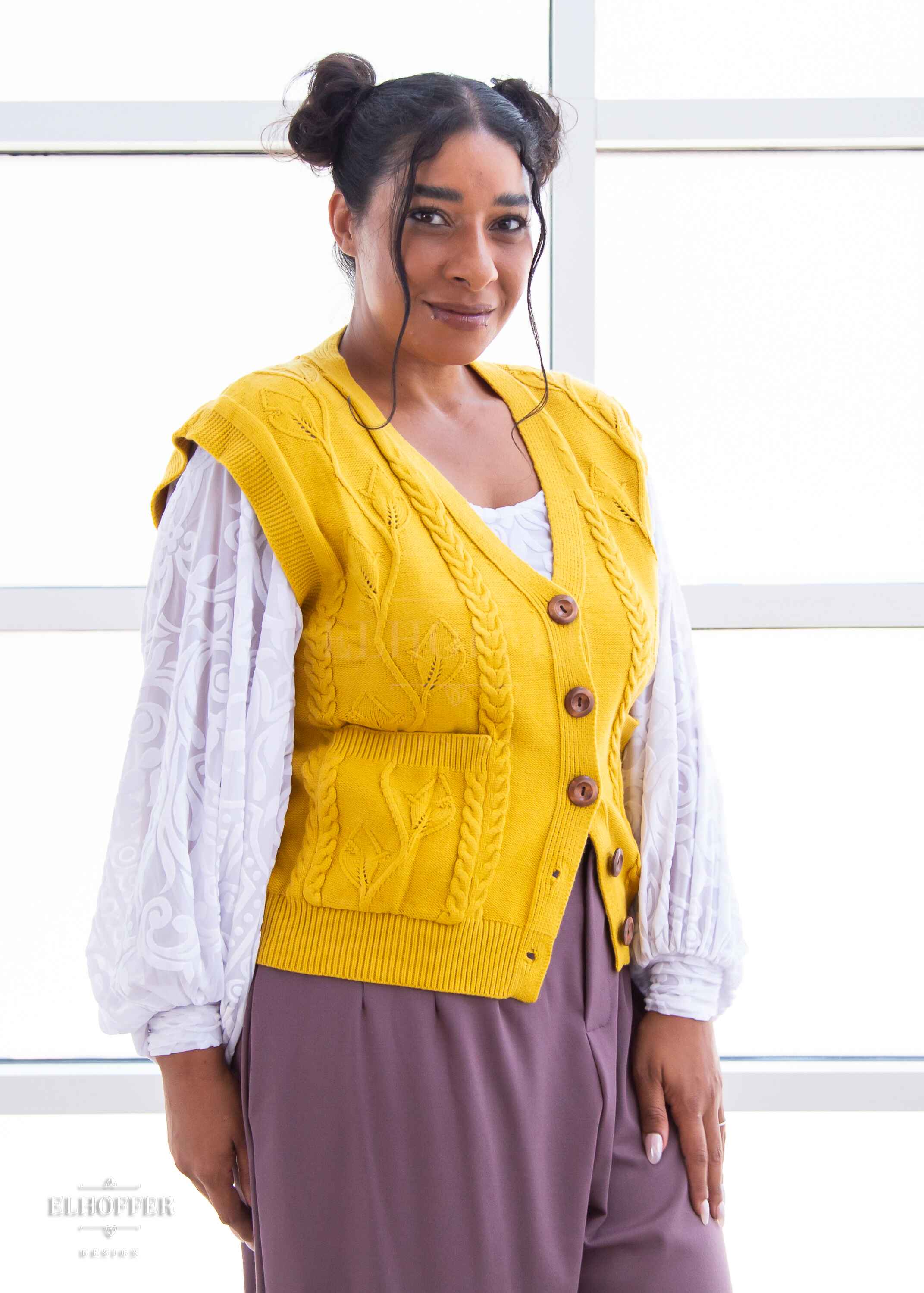 Janae, a light brown skinned L model with dark brown hair in space buns, is wearing a golden yellow button up knit vest with a leafy vine and cable knit pattern, light brown buttons, and front pockets. She paired the vest with a white burnout velvet top and mauve trousers.