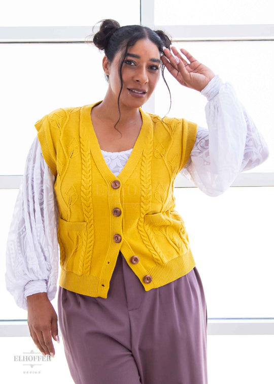 Janae, a light brown skinned L model with dark brown hair in space buns, is wearing a golden yellow button up knit vest with a leafy vine and cable knit pattern, light brown buttons, and front pockets. She paired the vest with a white burnout velvet top and mauve trousers.