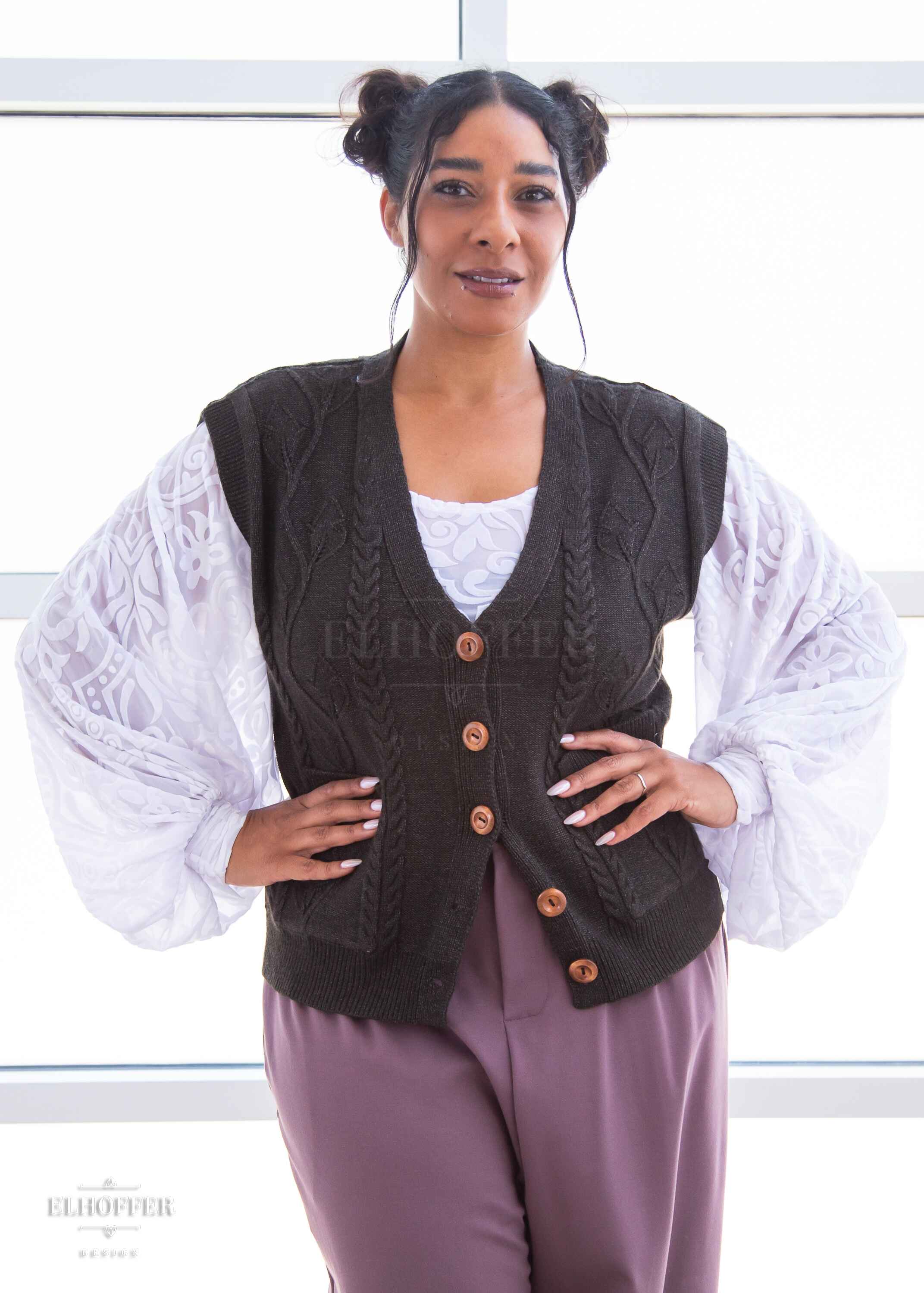 Janae, a light brown skinned L model with dark hair in space buns, is wearing the M (L-2XL) sample of a dark grey button up knit vest with a leafy vine and cable knit pattern, light brown buttons, and front pockets over a white long sleeve burnout velvet top with billowing sleeves and mauve pleated trousers.