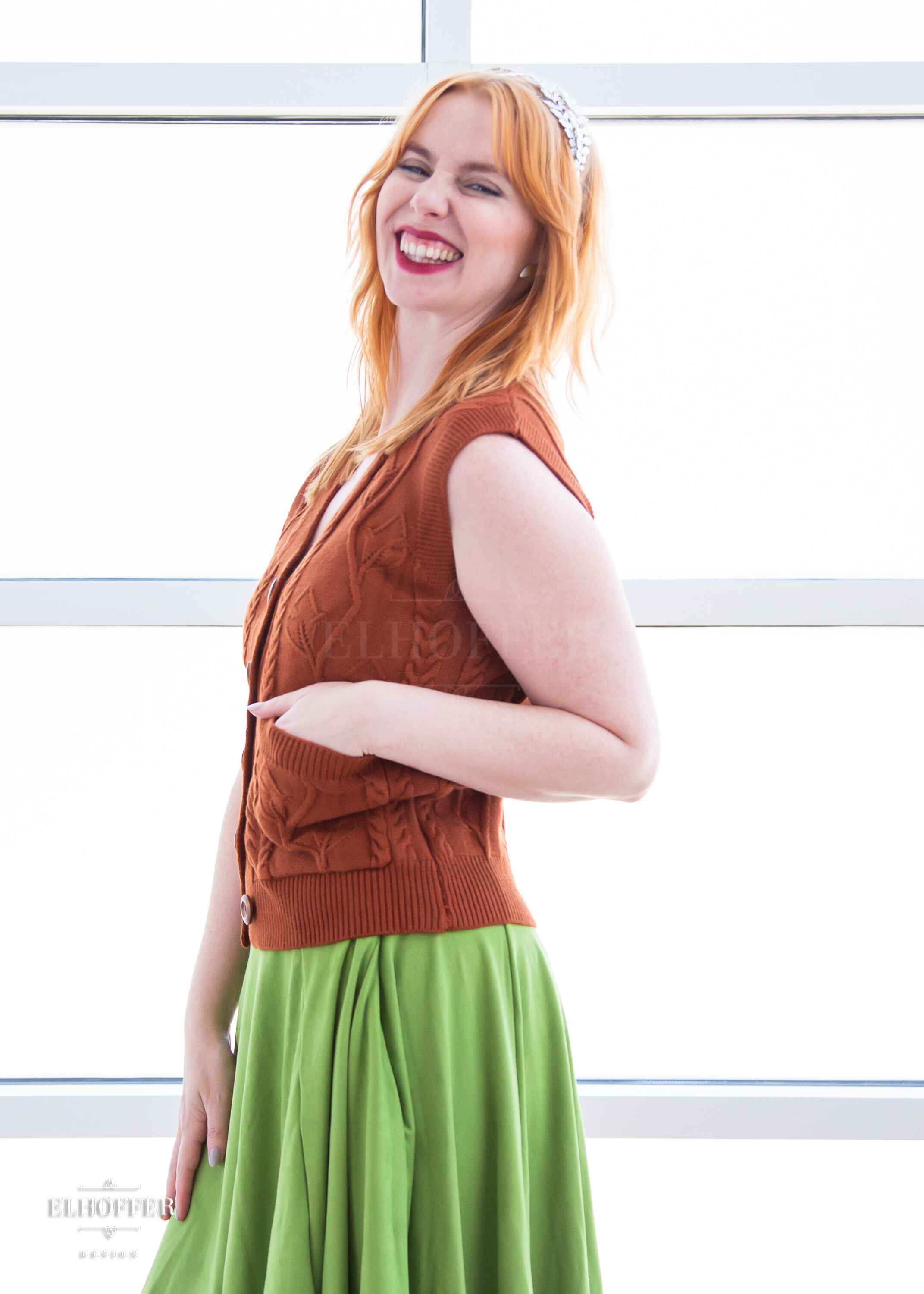 A side view of Harley, a fair skinned S model with shoulder length strawberry blonde hair, smiling while wearing a pumpkin orange button up knit vest with a leafy vine and cable knit pattern, light brown buttons, and front pockets.