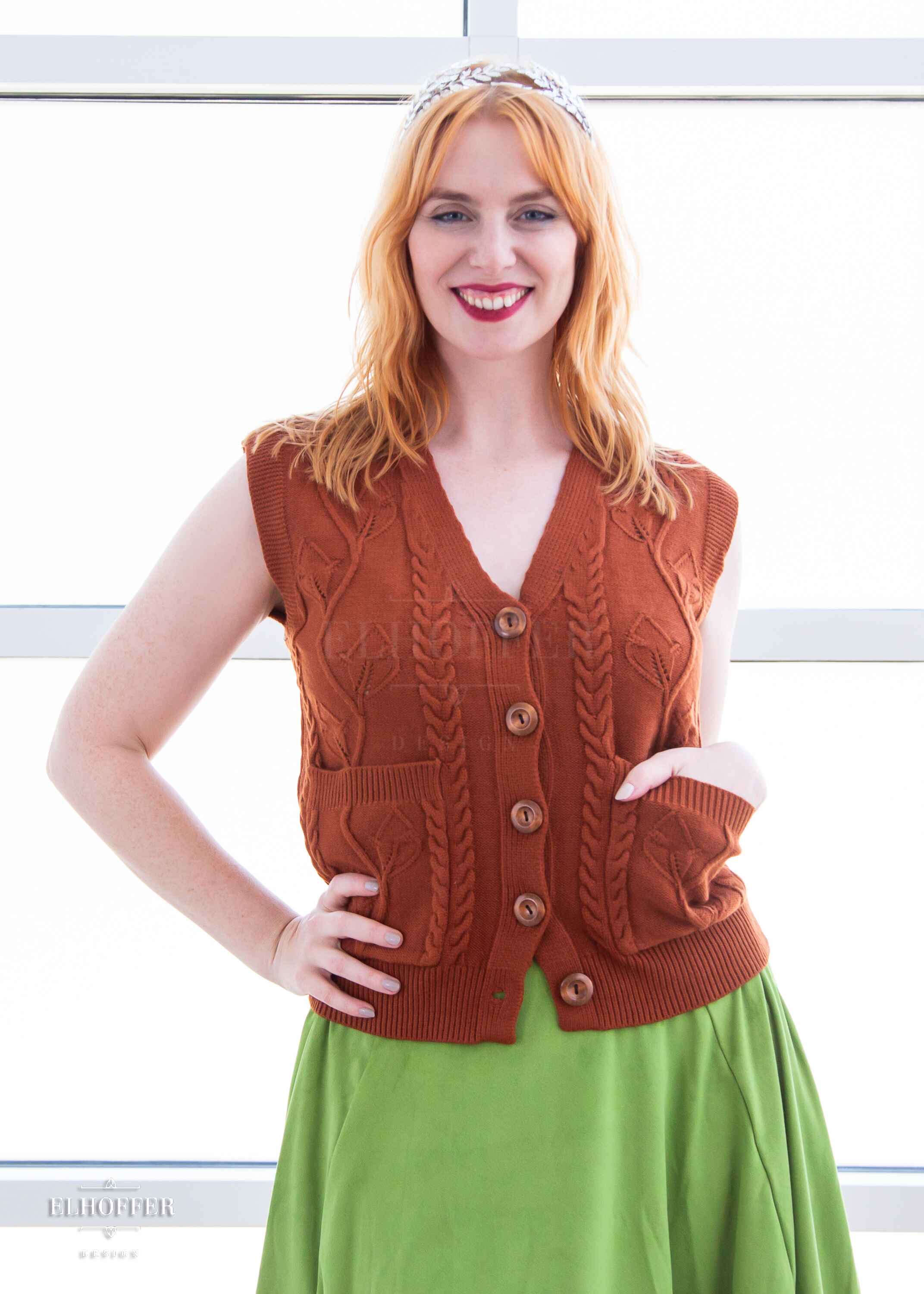 Harley, a fair skinned S model with shoulder length strawberry blonde hair, is smiling while wearing a pumpkin orange button up knit vest with a leafy vine and cable knit pattern, light brown buttons, and front pockets.