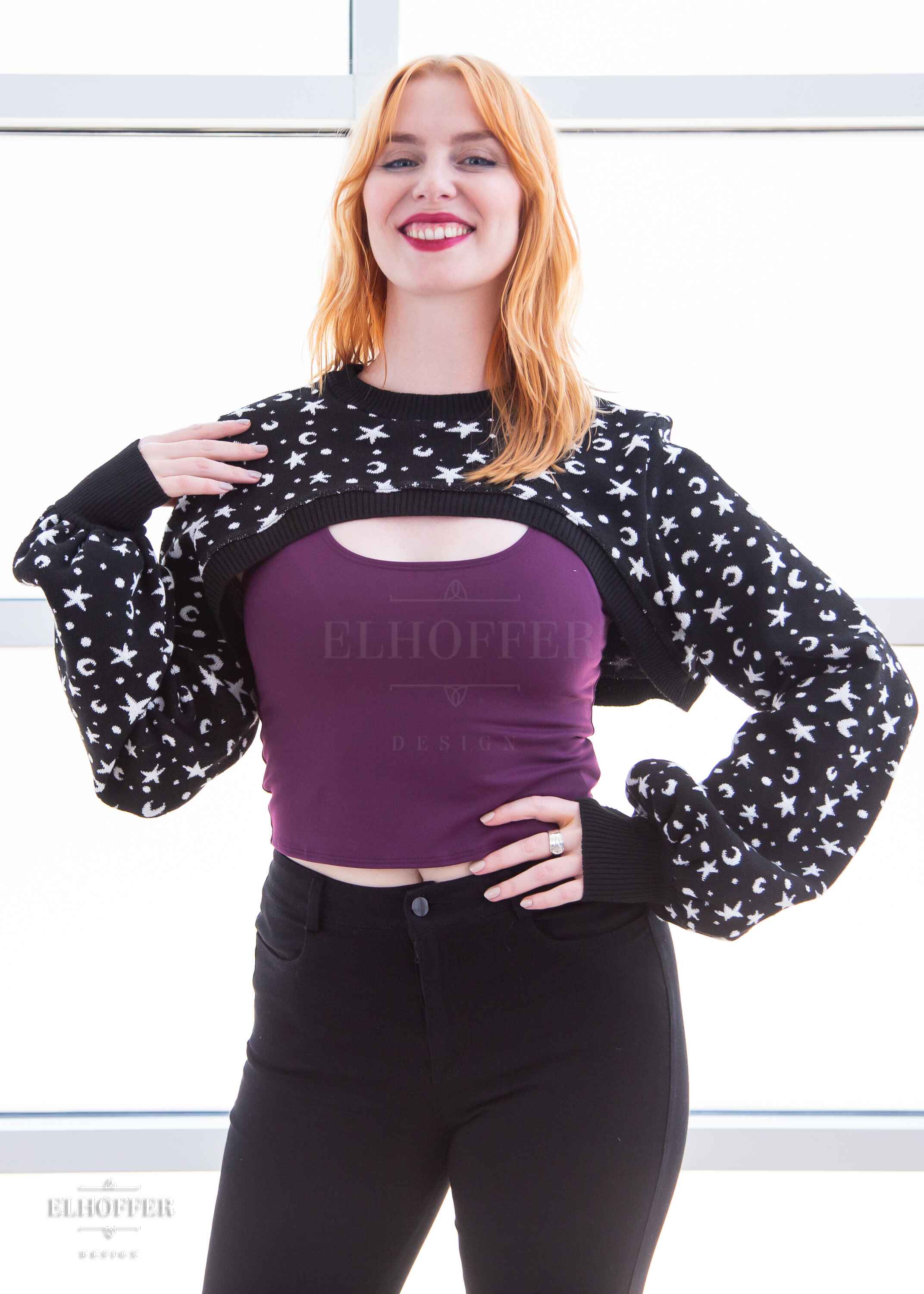 Harley, a fair skinned S model with shoulder length strawberry blond hair, is smiling while wearing the L sample of a black crew neck super cropped knit shrug sweater with a white star and moon pattern, long billowing sleeves and thumbholes. She would order the S for a slightly less oversize look.