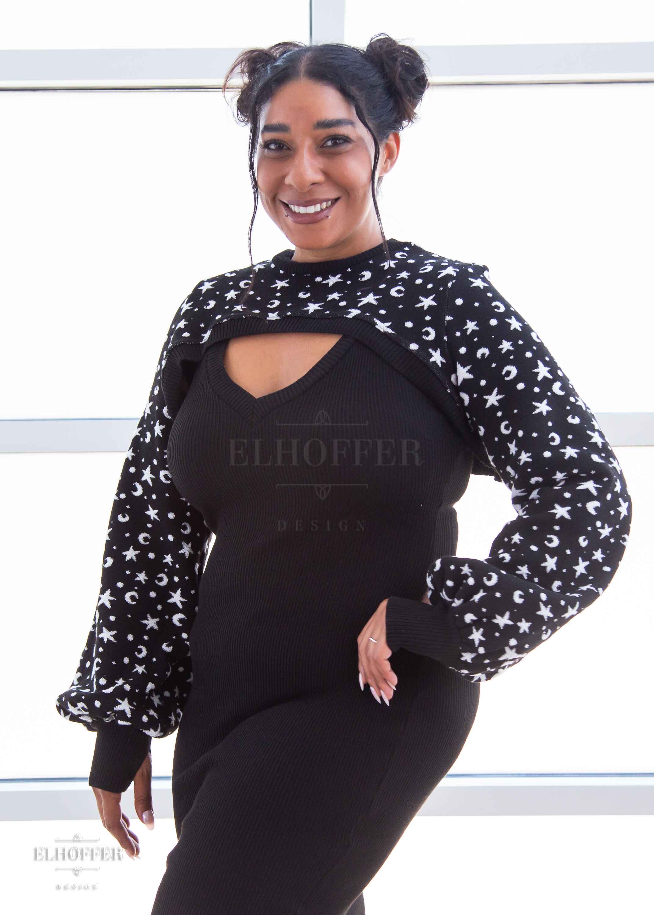 Janae, a light brown skinned L model with dark hair in space buns, is smiling while wearing a black super cropped crew neck knit shrug sweater with a white star and moon pattern, long billowing sleeves, and thumbholes.